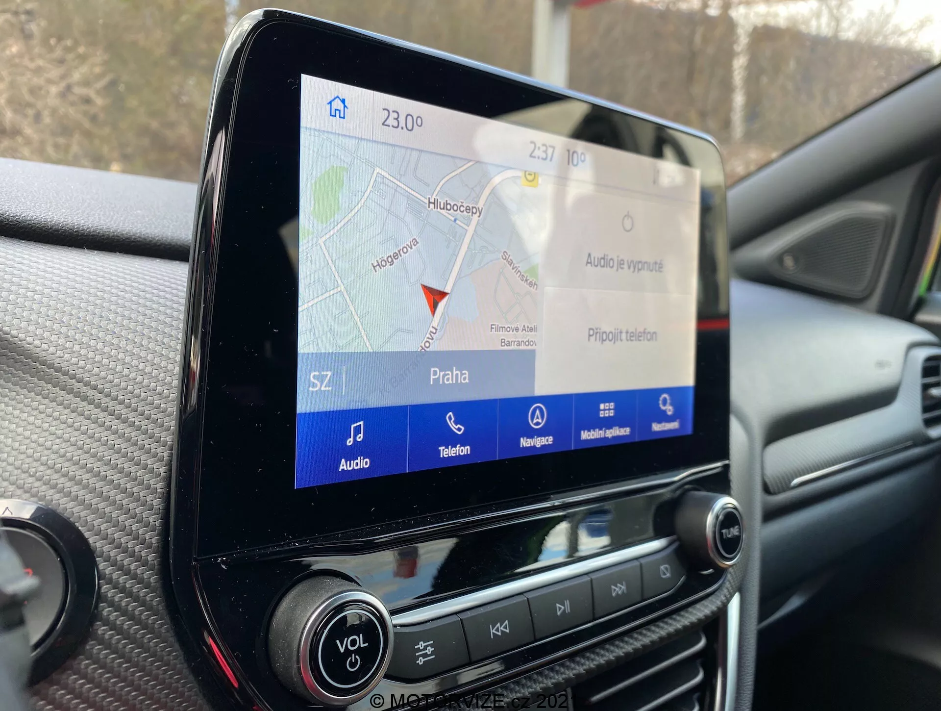 Närbild av infotainment pekskärmen i en Ford Puma 2020, som visar GPS-navigeringskartan märkt 'Praha', med ikoner för ljud-, telefon- och navigeringsfunktioner, omgiven av fysiska kontrollknappar och vred på instrumentbrädan.
