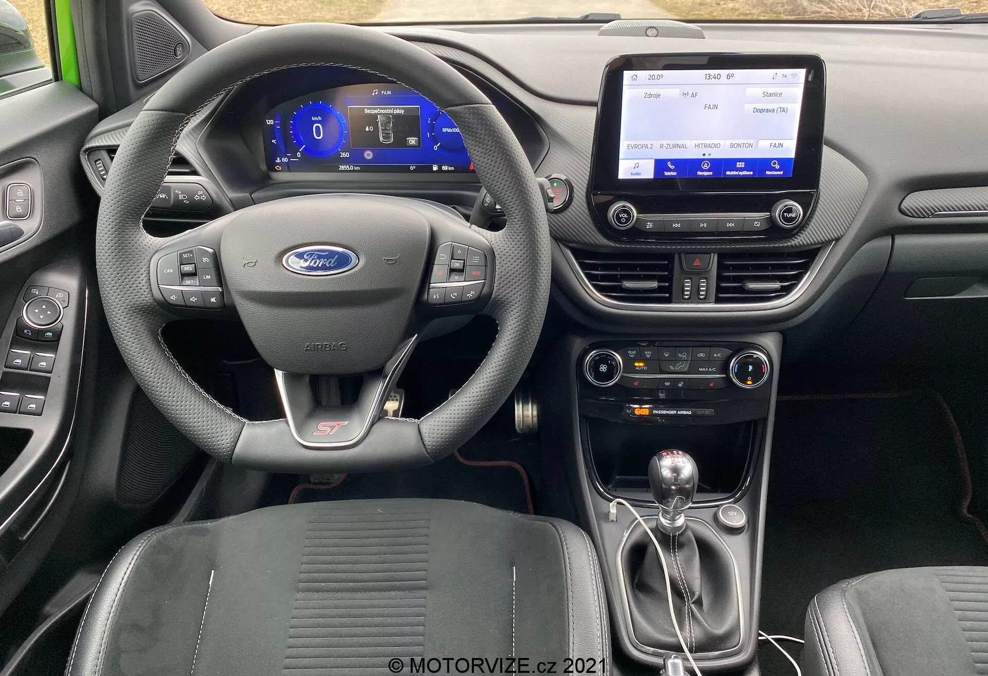 Interiörbild av en Ford Puma ST 2020 från bakre sätesområdet, som framhäver läderklädd ratt med Ford-logotyp och kontrollknappar, digitalt instrumentkluster med blå bakgrundsbelysning, central infotainmentskärm, modern mittkonsol med klimatanläggningskontroller, manuell växelspak med läderöverdrag och metalliska accenter, formade sportstolar samt högkvalitativa material inklusive läder, plast och metalliska accenter.