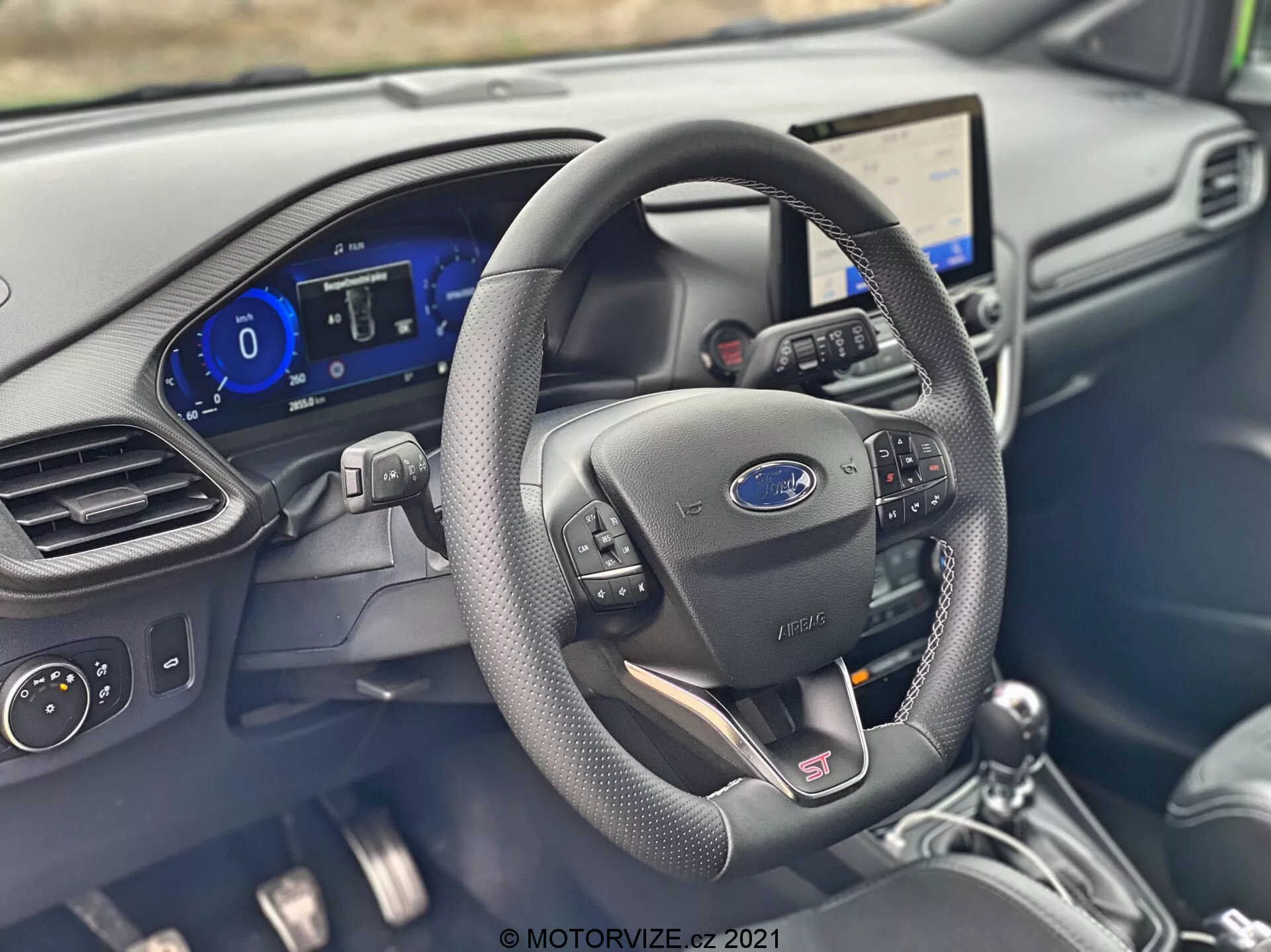 Interiörbild av 2020 Ford Puma ST som visar förarens cockpit från framsätets passagerarperspektiv, med en svart läderklädd multifunktionsratt med Ford- och 'ST'-emblem. Noterbara detaljer innefattar en digital instrumentpanel som visar hastighetsmätare, kraft- och laddningsmätare, samt ett vinklingsbart infotainmentsystem med pekskärm och uppkopplingsfunktioner. Interiören har en modern design med mörka färgscheman, lädertexturerade plaster och metalliska accenter samt klimatkontrollgränssnitt, vilket understryker bilens avancerade teknik och prestandainriktning.
