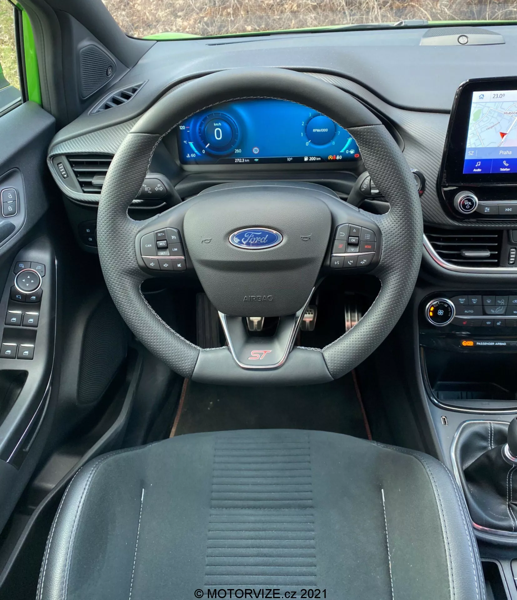 Interiörbild från förarens perspektiv av en Ford Puma ST 2020 som visar en modern och välutrustad kupé. Ratten med Ford-logotypen har inbyggda kontroller för ljud- och farthållare, och ST-märket längst ner indikerar en sportig trim. Bakom ratten finns en helt digital instrumentpanel som visar hastighet och bränslenivå på en blåtonad skärm. Mittkonsolen till höger innehåller ett infotainmentsystem med pekskärm som visar en navigationskarta, tillsammans med prydligt organiserade klimatkontroller som antyder dubbla zoner. De svarta, eleganta materialen och den samtida förarfokuserade designen är uppenbara, med hållbar klädsel synlig på sätena. Bilden ger en ögonhöjds vy från förarsätet, fokuserar på fordonets avancerade digitala gränssnitt och komfortfunktioner.