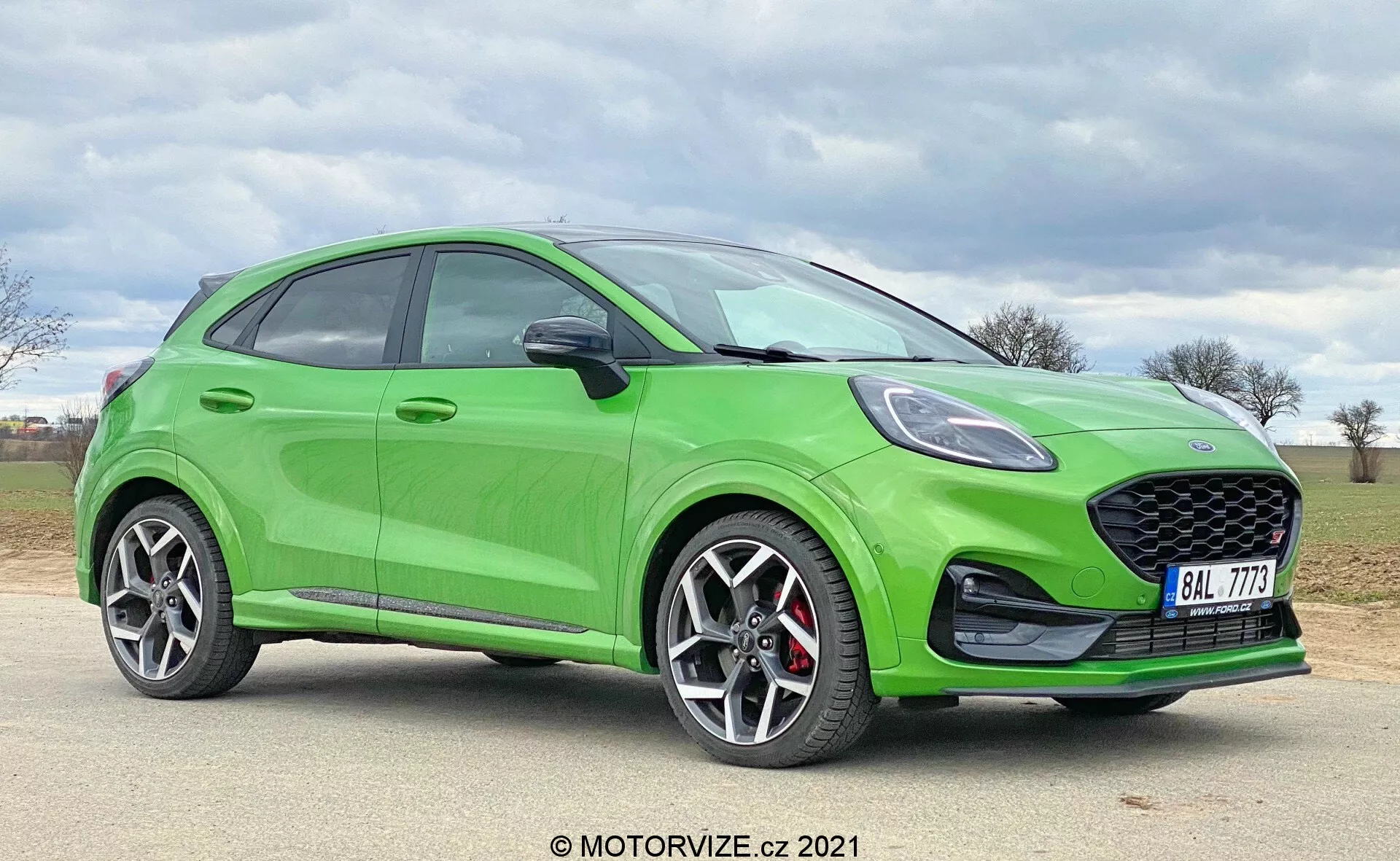 En ljusgrön Ford Puma ST 2020 hatchback fotograferad från en sidovinkel, som visar bilens profil från fronten till bakdelen. Den lågvinklade bilden framhäver den framträdande bikakegrillen, sportiga strålkastare, konturerad motorhuv och stora lättmetallfälgar med röda bromsok. Den eleganta, aerodynamiska designen inkluderar en gradvis sluttande taklinje, synliga karaktärslinjer och ett uns av en bakre spoiler.