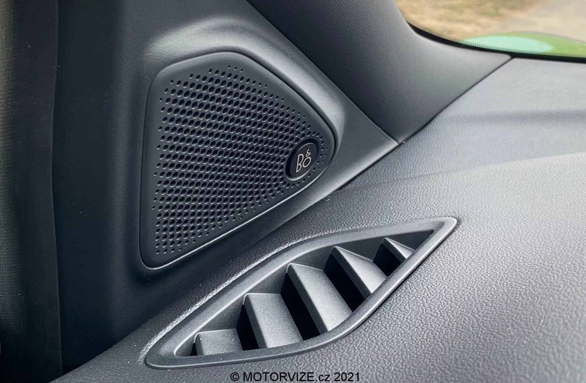 Närbild av Ford Puma ST 2020 interiör, med fokus på ett högkvalitativt högtalarsystem från Bang & Olufsen med ett honungskakemönstrat galler och en justerbar luftventil nedanför. Bilden fångar de texturala detaljerna i högtalaren och ventilationssystemet i en lätt sned vinkel, vilket betonar lyxbilsfunktioner och avancerad bildesign.