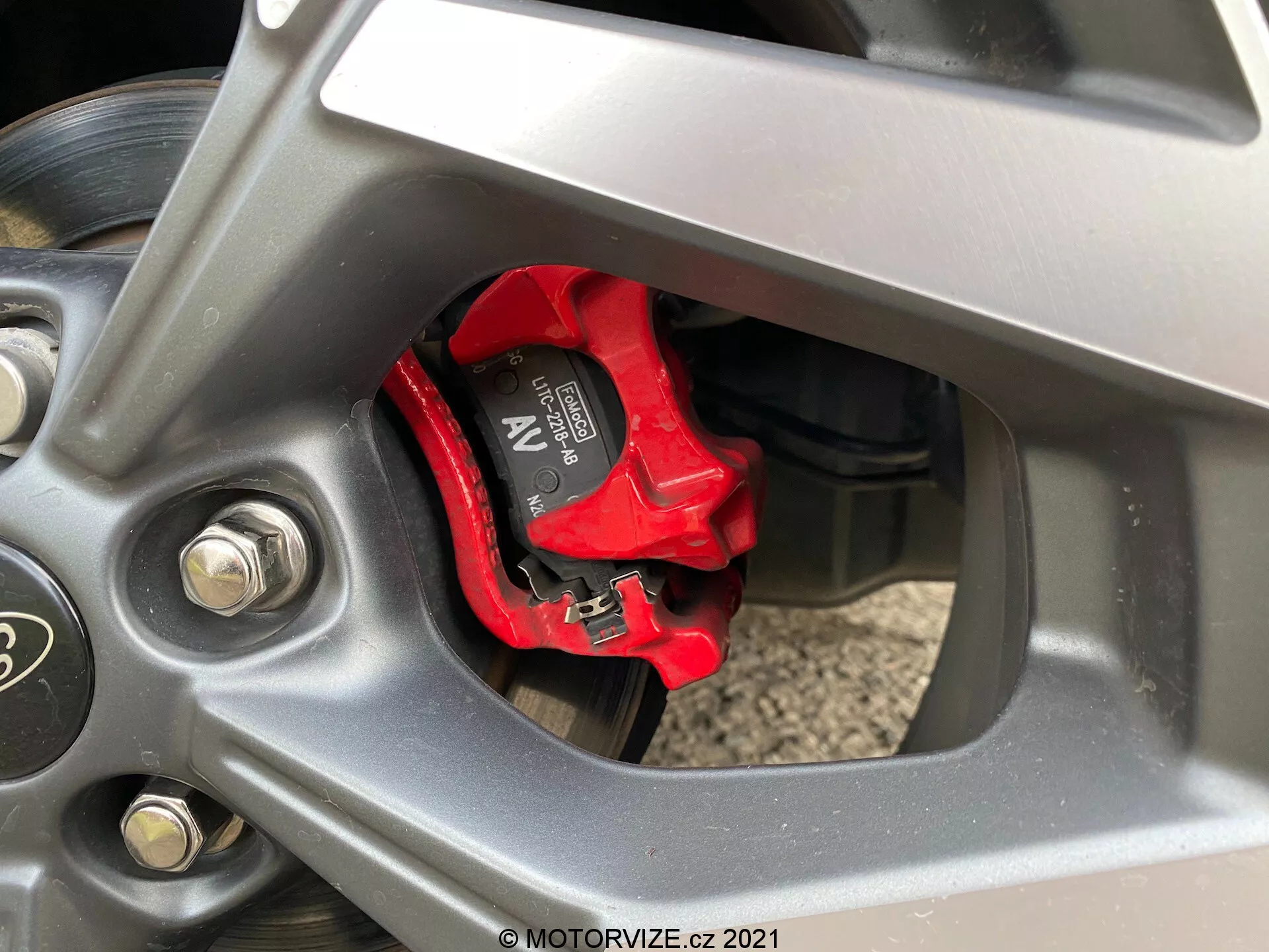 Detaljerad närbild av ett Ford Puma ST 2020 fordonshjul, fokuserat på den högpresterande Brembo bromsoket i ljusröd och ventilerad bromsskiva. Hjulet med fem delade ekrar har en mörk metallisk finish, med synliga silverfärgade hjulmuttrar och delvis synlig sidovägg på däcket. Suspension komponenter svagt synliga i bakgrunden.