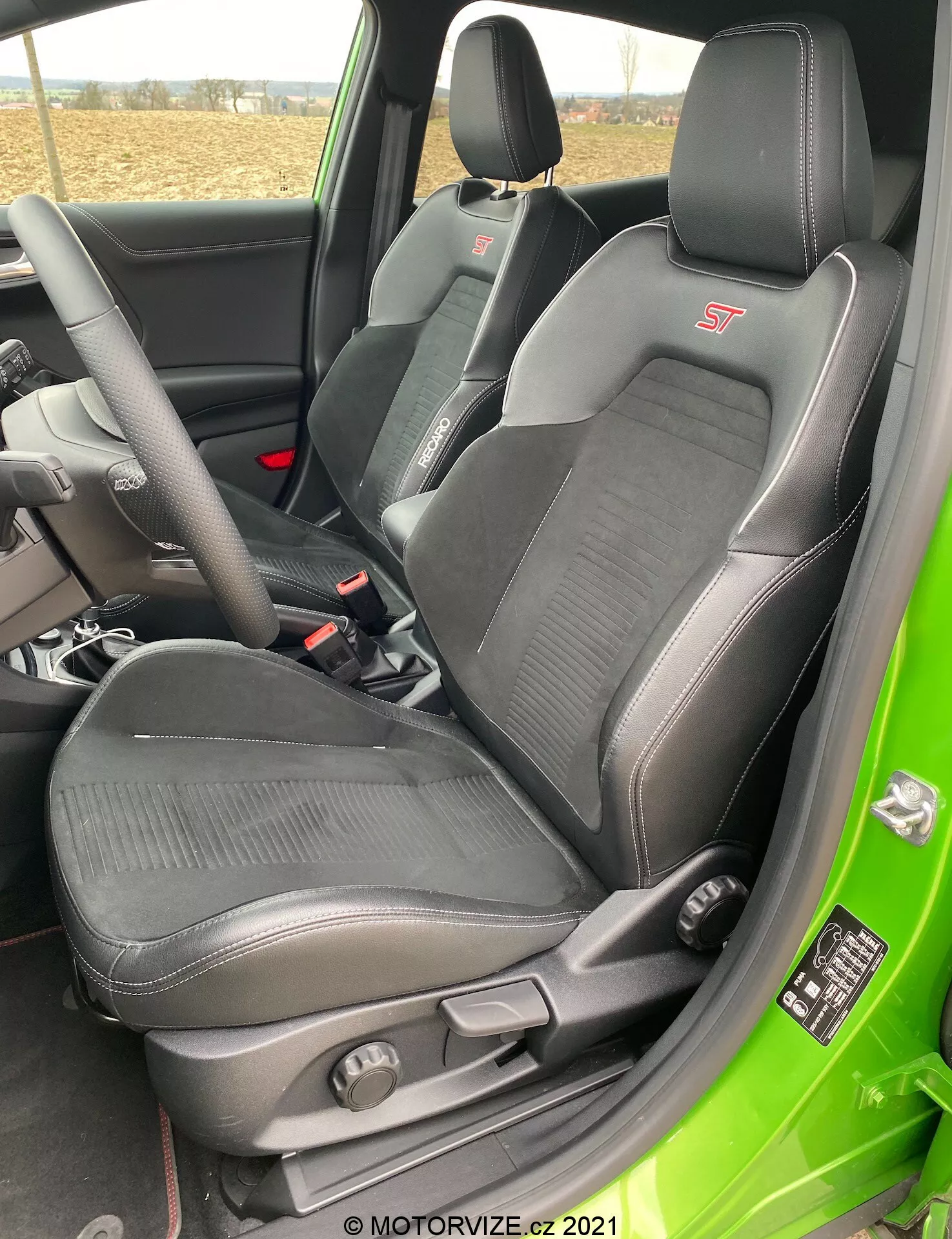 Interiörbild av en Ford Puma ST 2020, tagen från den öppna förarsidan med fokus på den sportiga förarstolen. Stolen har ett integrerat nackstöd, framträdande sidostöd och lårstöd samt en klädsel i en kombination av läder och tyg med mönstrade tygsektioner. En 'ST'-logotyp är broderad på den övre delen av stolen. Manuella justeringsspakar och en roterande ratt för svankstöd är synliga. I bakgrunden syns delvis ratten och säkerhetsbältet, där det naturliga dagsljuset lyser upp interiören.
