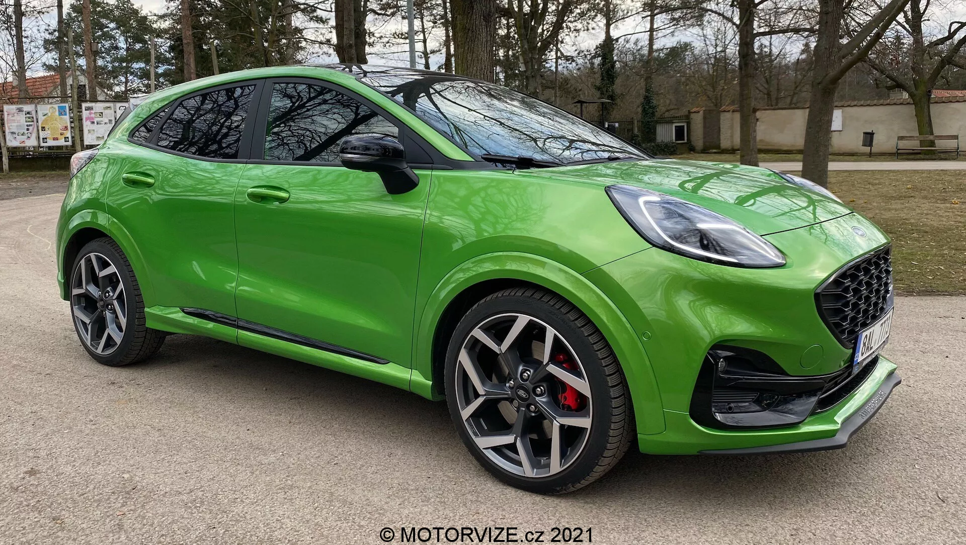 En ljusgrön Ford Puma ST 2020 fotograferad från en låg trekvarts vinkel framifrån, som framhäver dess eleganta och sportiga design med ett bikakemönstrat grill, skarpa LED-strålkastare, mörka flerspoked aluminiumhjul, röda bromsok, skulpterad sidoprofil, speglar med integrerade blinkers, sluttande taklinje, blank lack och ytterligare aerodynamisk exteriörtrimning. Bilen är parkerad utomhus med träd och en byggnad ur fokus i bakgrunden.