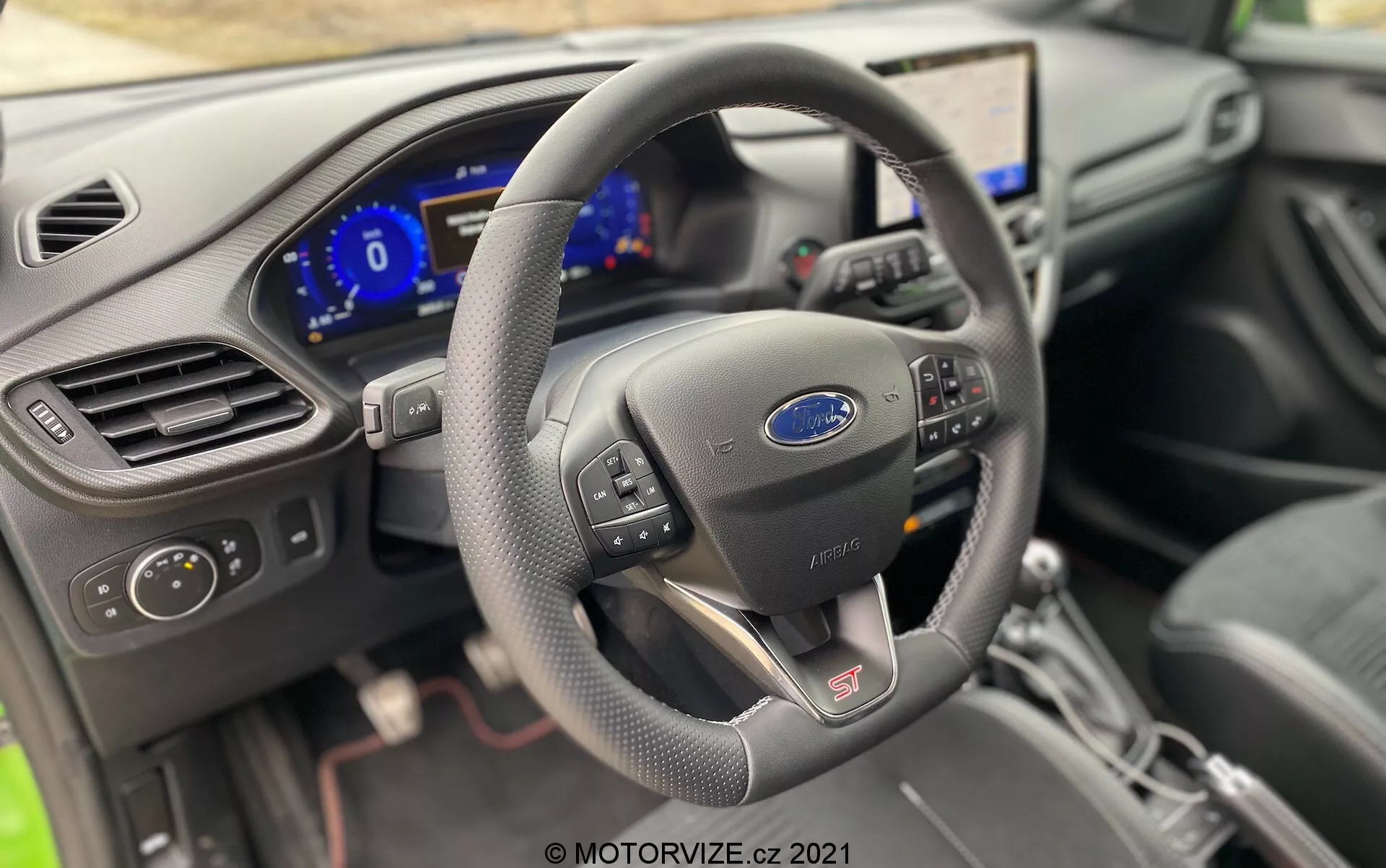 Interiörvy av förarområdet i 2020 Ford Puma ST från en lätt vänstervinkel, med fokus på en läderklädd multifunktionell ratt med kontrollknappar, en digital instrumentpanel som visar fordonsinformation, en mittkonsol med en stor pekskärm för infotainment, synlig luftventil och strålkastarkontrollknapp, en central växelspak med läderskydd och S-symbol som möjligen indikerar sportläge, samt kanten av en mörkt klädd stol, vilket visar den teknologiskt avancerade och moderna designen av bilens interiör.