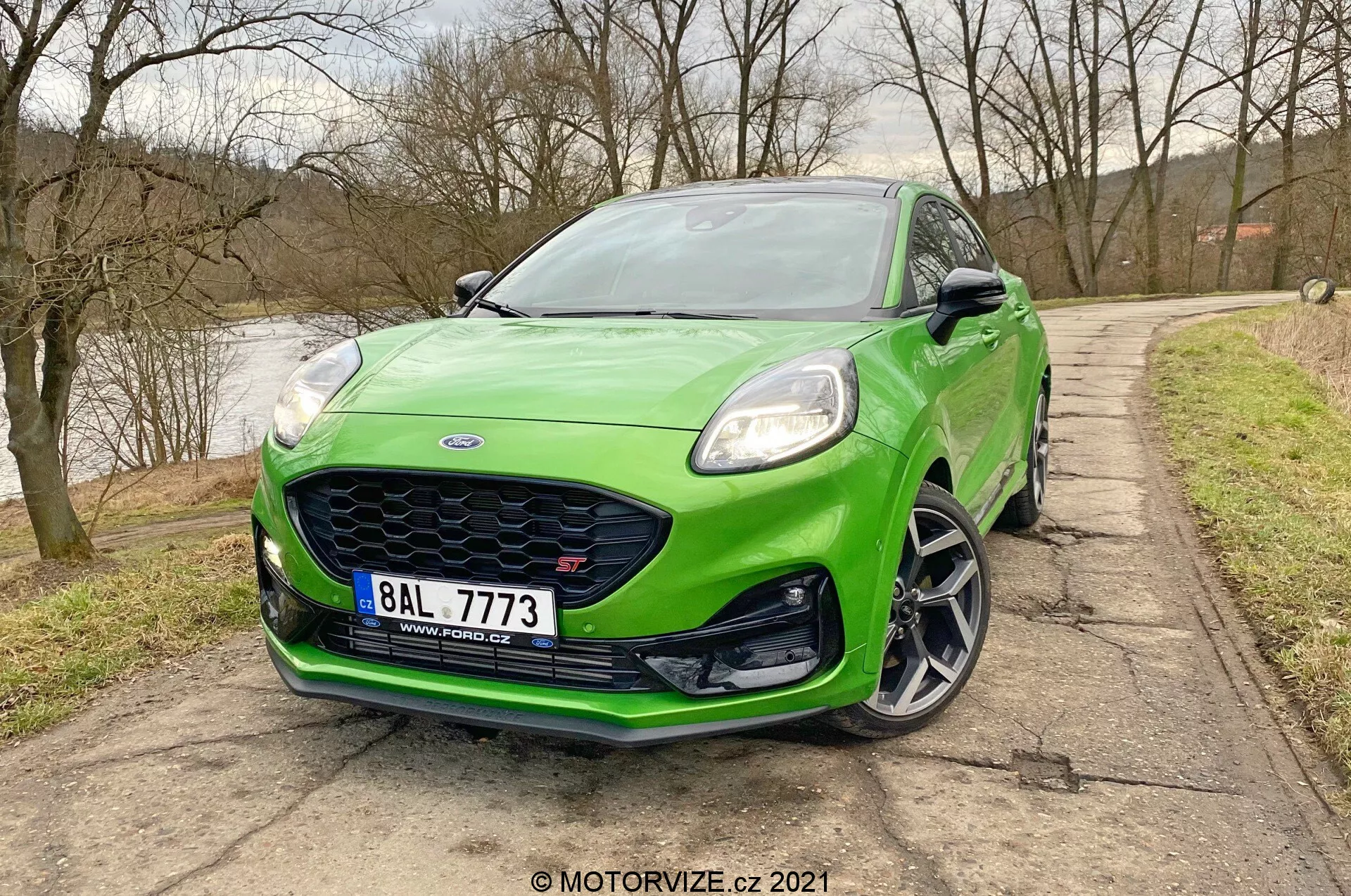 Trekvartsfrontal vy av en ljusgrön Ford Puma ST 2020 halvkombi, som visar upp dess frontdesignaspekter inklusive vänster strålkastare, främre stötfångare, grill och konturen av vänster skärm. Den smidiga motorhuven och vindrutan är också synliga. Bilden är tagen i en lätt nedåtgående vinkel, vilket framhäver den aggressiva främre stötfångaren med luftintag och splitter, eleganta LED-strålkastare, mångekrade hjul med lågprofildäck och en integrerad blinkers i sidospegeln. Närbilden betonar fordonets sportiga och moderna estetik.