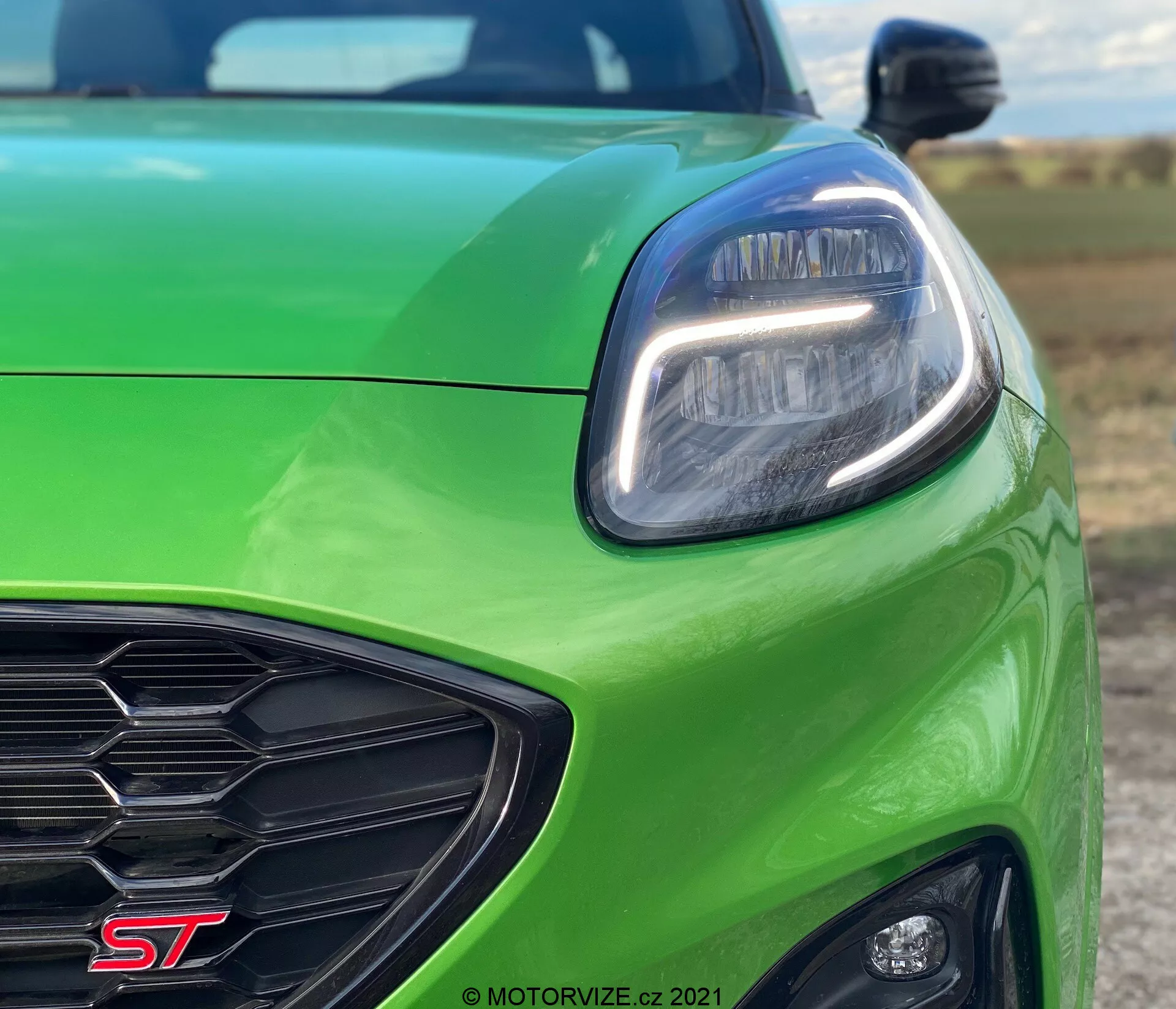 Närbild av vänster främre fjärdedel av en grön Ford Puma ST 2020. Bilden betonar den moderna LED-strålkastaren, det mörka främre gallret med en ST-märke, den eleganta främre stötfångaren och den glansiga huven med en sportig konturlinje. Den låga vinkeln framhäver bilens aerodynamiska design och prestandafunktioner.