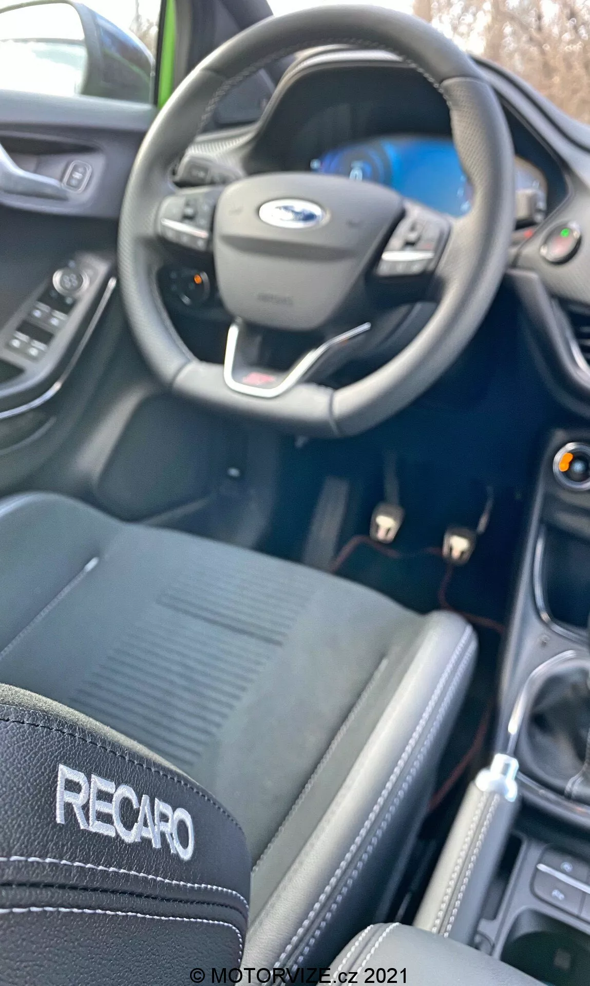 Interiörvy av en Ford Puma ST 2020, som visar den treekrade ratten med inbyggda kontroller och Ford-logotyp; delvis synlig mätargrupp på en modern instrumentpanel med kurvor. Den centrala infotainment-skärmen antyds subtilt. Ett svart 'RECARO'-märke på en stol med synliga sömmar indikerar en högprestandainriktning, och en manuell växelspak i en panel med metallutseende tyder på manuella växlingsmöjligheter. Den mjuka fokusen i bilden ger en spontan känsla, som betonar sportiga och prestandarelaterade attribut.