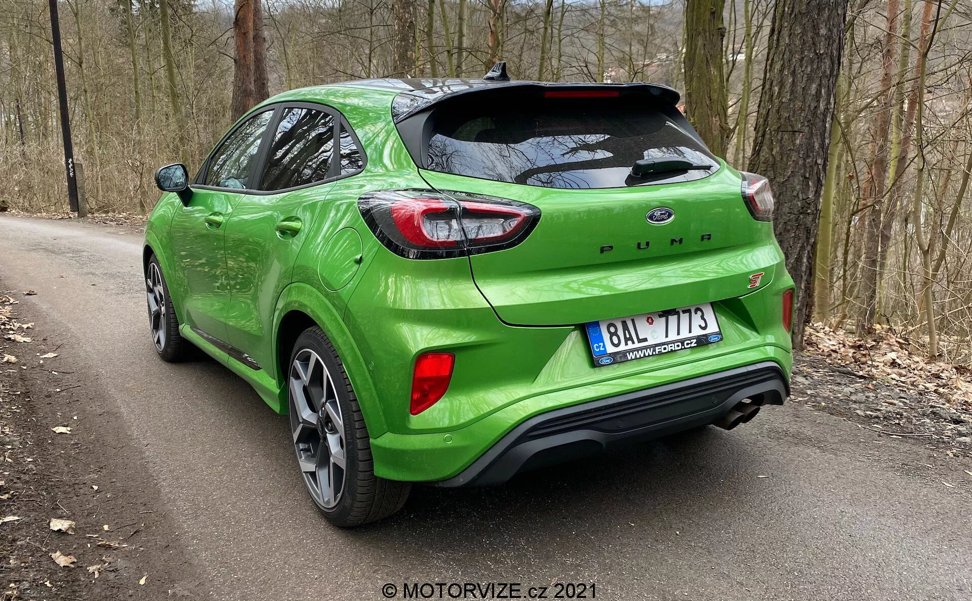 Trekvarts bakåtvänd vy av den gröna Ford Puma 2020-modellen som visar passagerarsidan och bakre designen med synlig bakluckespoiler, LED-baklyktor, Ford-logotyp, Puma-namnskylt, lättmetallfälgar, parkeringssensorer integrerade i stötfångaren och avgasrör på högersidan. Positionerad på en skogsväg, vilket framhäver en utomhus- eller äventyrsmiljö.