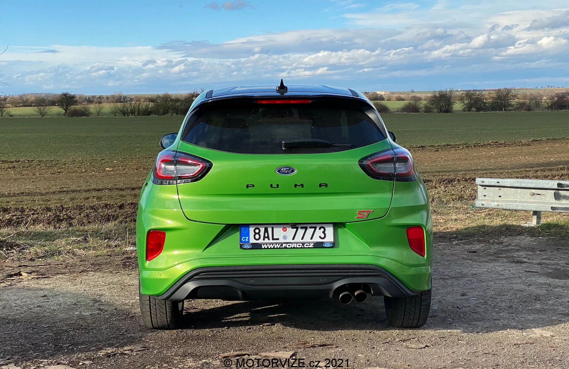 Bakre vy av en ljusgrön 2020 Ford Puma ST, fotograferad från marknivå med fokus på designfunktionerna på bilens baksida. Bilden visar hela bilens bredd mot en klar himmel och ett öppet landskap, vilket tyder på landsbygdsomgivning eller utkanten av ett stadsområde. Högdagarna inkluderar en modern hatchback-design, bakspoiler, hajfensantenn, moderna bakljus som omsluter sidorna, distinkt bakre stötfångare med parkeringssensorer, dubbla avgasrör, backkamera under registreringsskylten och elektronisk bagagelucka.