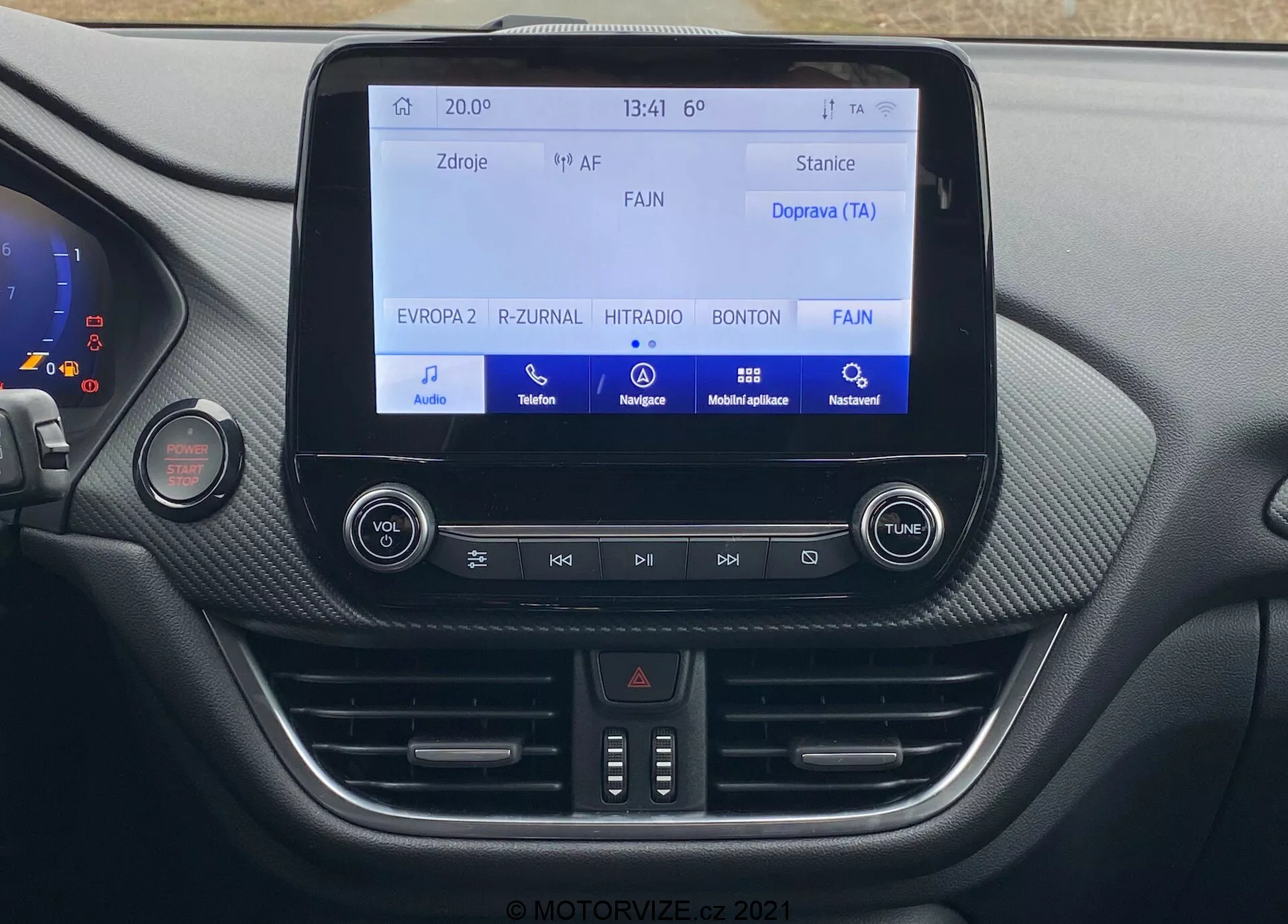 En detaljerad vy av den centrala instrumentpanelen i en Ford Puma 2020 (Puma-ST), med fokus på infotainmentsystemet och kontroller från passagerarsätets perspektiv. Den centrala pekskärmen visar ljud- och mediefunktioner, medan fysiska knappar och temperaturkontroller för den tvåzoniga klimatkontrollen syns nedanför. Den moderna designen inkluderar en blandning av texturer och finish med ett mjukt material och ett kolfiberliknande mönster.
