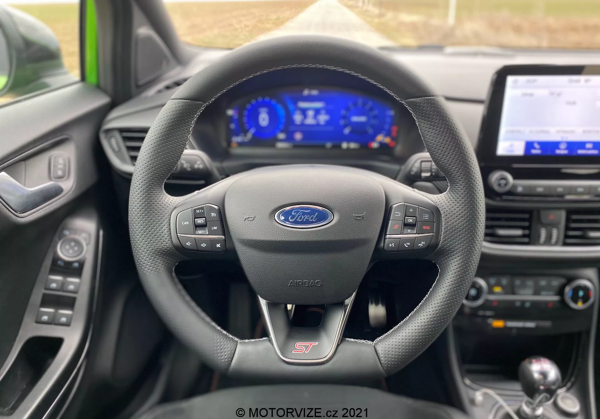 Förarperspektiv av interiören i en Ford Puma 2020 (Puma-ST), med fokus på den läderklädda multifunktionsratten med Ford-logo, ST-logotyp och digital instrumentpanel som visar hastighet, varvtal och bränslenivå. Mittkonsolen med infotainmentskärm, klimatanläggningens kontroller och manuell växelspak är också synliga, upplysta med en sval blå ton.