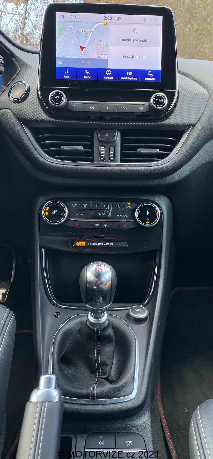 Närbild med vertikal vy av mittkonsolen i Ford Puma 2020, som visar infotainmentskärmen med en karta, klimatkontroller och manuell växelspak med ytterligare körlägesreglage. Konsolen har en blandning av mjukplast och läder med kontrastsömmar, vilket betonar bilens interiörkvalitet och ergonomi.