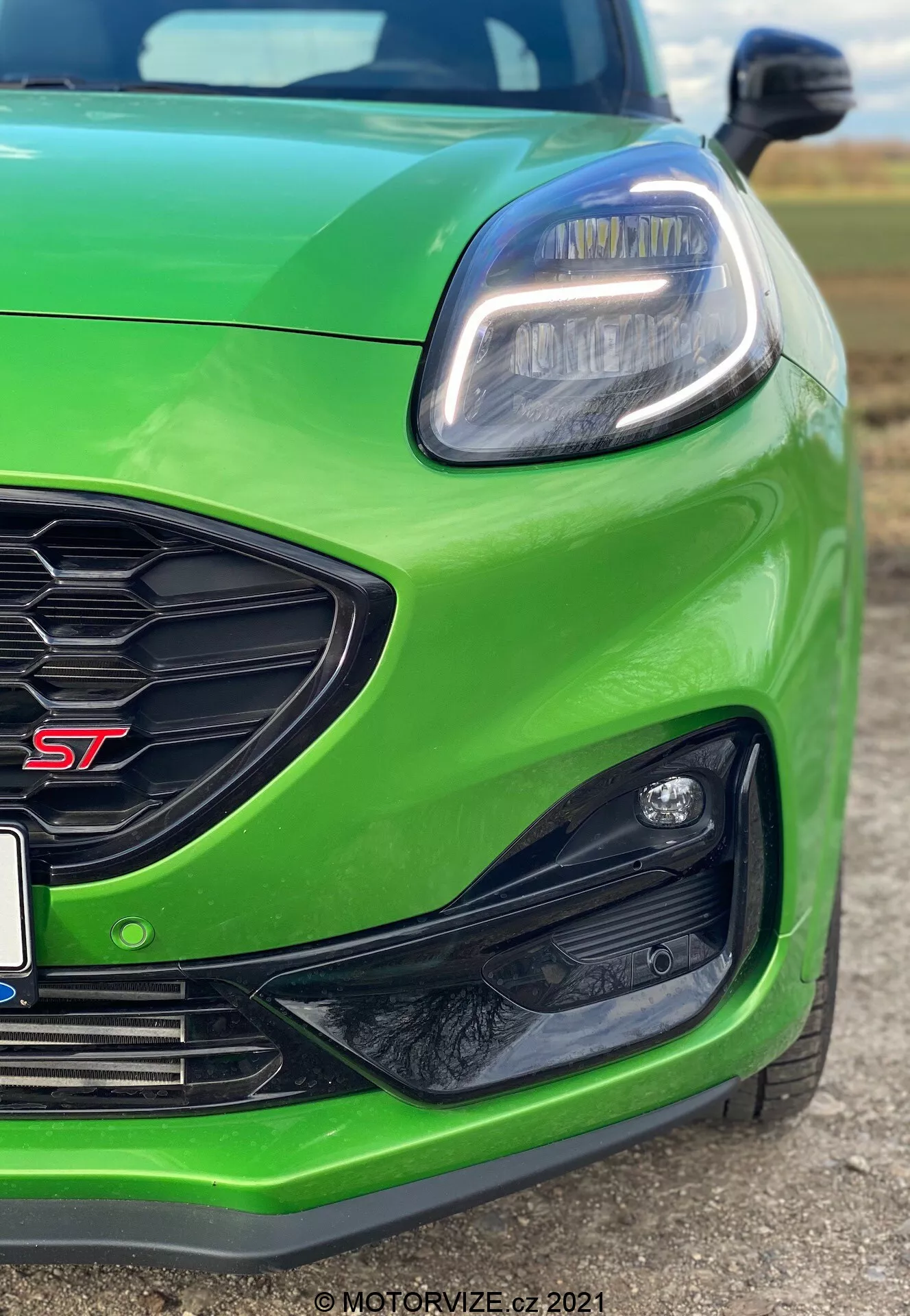 Närbild av den vänstra främre delen av en ljusgrön Ford Puma ST 2020, där den vänstra LED-strålkastaren med modern, kantig design och LED-dagbelysning syns tydligt, tillsammans med ett honungsmönstrat galler med en röd 'ST'-märkning och en kantig svart infattning på främre stötfångaren, troligtvis för dimljus eller sensorer. Den låga vinkeln betonar den sportiga hållningen och framhäver prestandainriktade egenskaper och tekniska framsteg i designen.