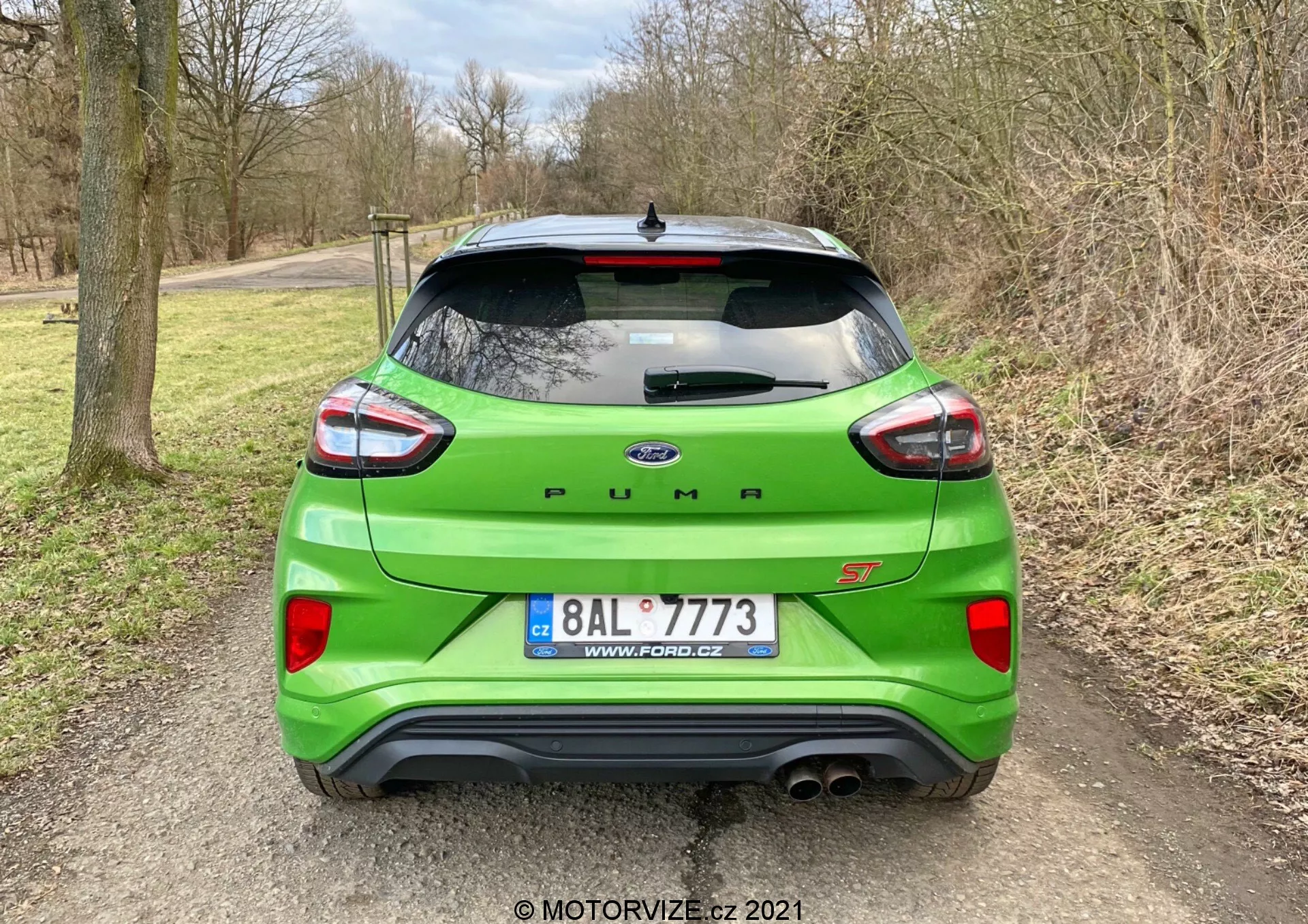 Bild av en ljusgrön Ford Puma ST tagen bakifrån, som visar dess sportiga designfunktioner inklusive en takspoiler, distinkta LED-baklyktor i bumerangform och dubbla avgasrör. Bilen markeras av en djärv 'PUMA'-text och 'ST'-emblem på bakluckan. Bakre stötfångaren integrerar parkeringssensorer, och ett mindre centralt galler under Ford-emblemet och registreringsskylten lägger till stil. Fordonet är parkerat på ett grus- eller jordområde, omgiven av träd och vegetation, vilket antyder en lantlig miljö.