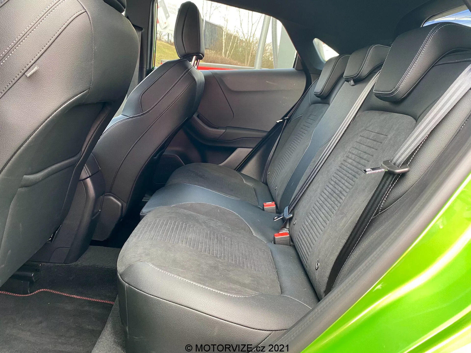 Närbild av interiören i Ford Puma 2020 (Puma-ST) visar baksätena från den högra sidan. Fokus ligger på den konturerade bakre soffan med en tvåfärgad design i läder och tyg. De mörka lädertexturerna kontrasterar med de ljusa tygpartierna med mönstrade inbuktningar. Synliga är justerbara nackstöd, röda säkerhetsbälten och högkvalitativa sömmar. Framsätet och del av dörrpanelen matchar sätesmaterialen, där framsätet visar förbättrat sidostöd.