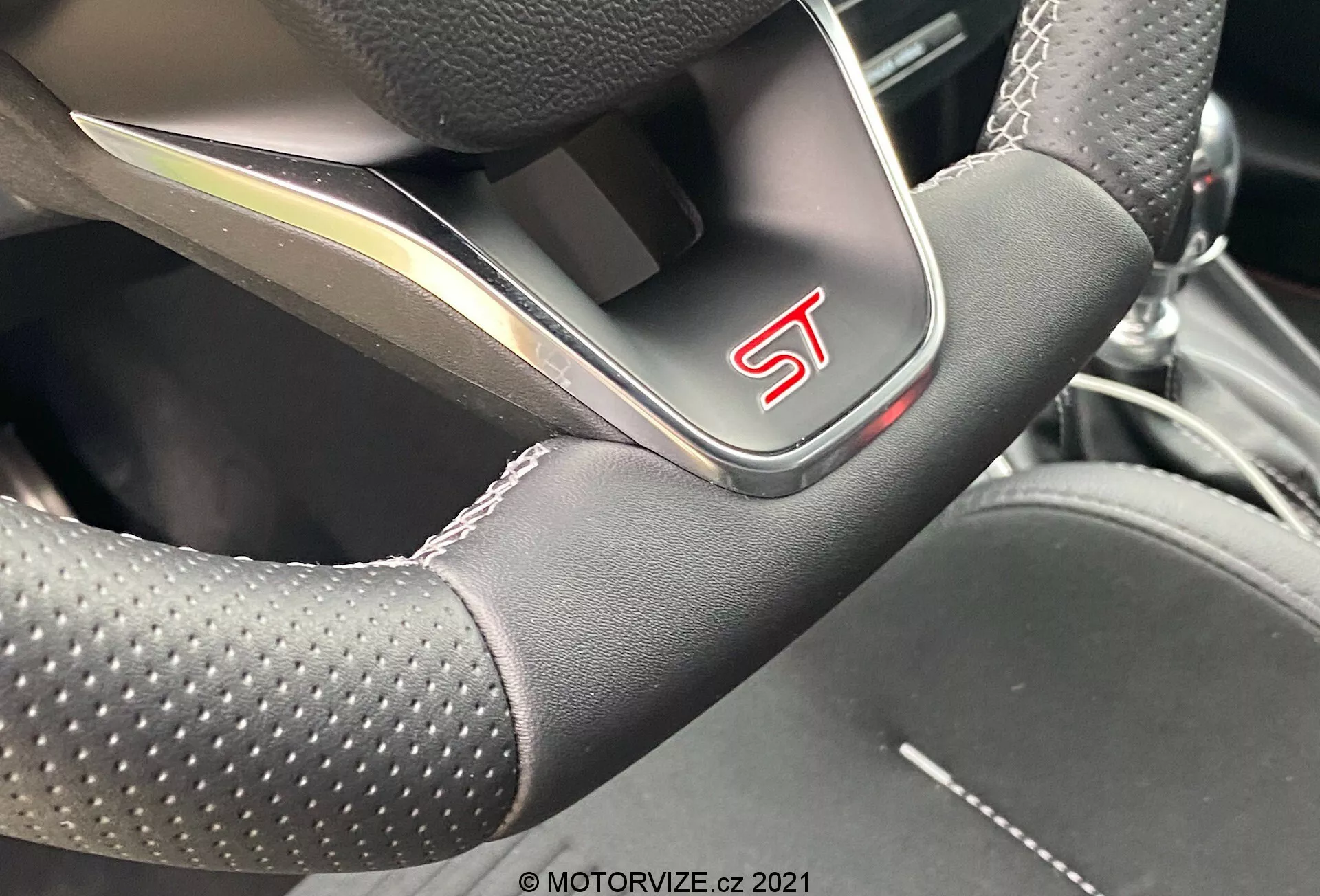 Denna närbild visar den nedre centrala delen av den sportiga ratten i en Ford Puma 2020 ST. De perforerade lädergreppen, platt botten, kontrastsömmar och metallisk trim framhäver dess premium- och prestandafokuserade egenskaper, med en synlig 'ST'-märkning. Vinkeln antyder att fotografen är placerad i förarsätet eller precis utanför förardörren.