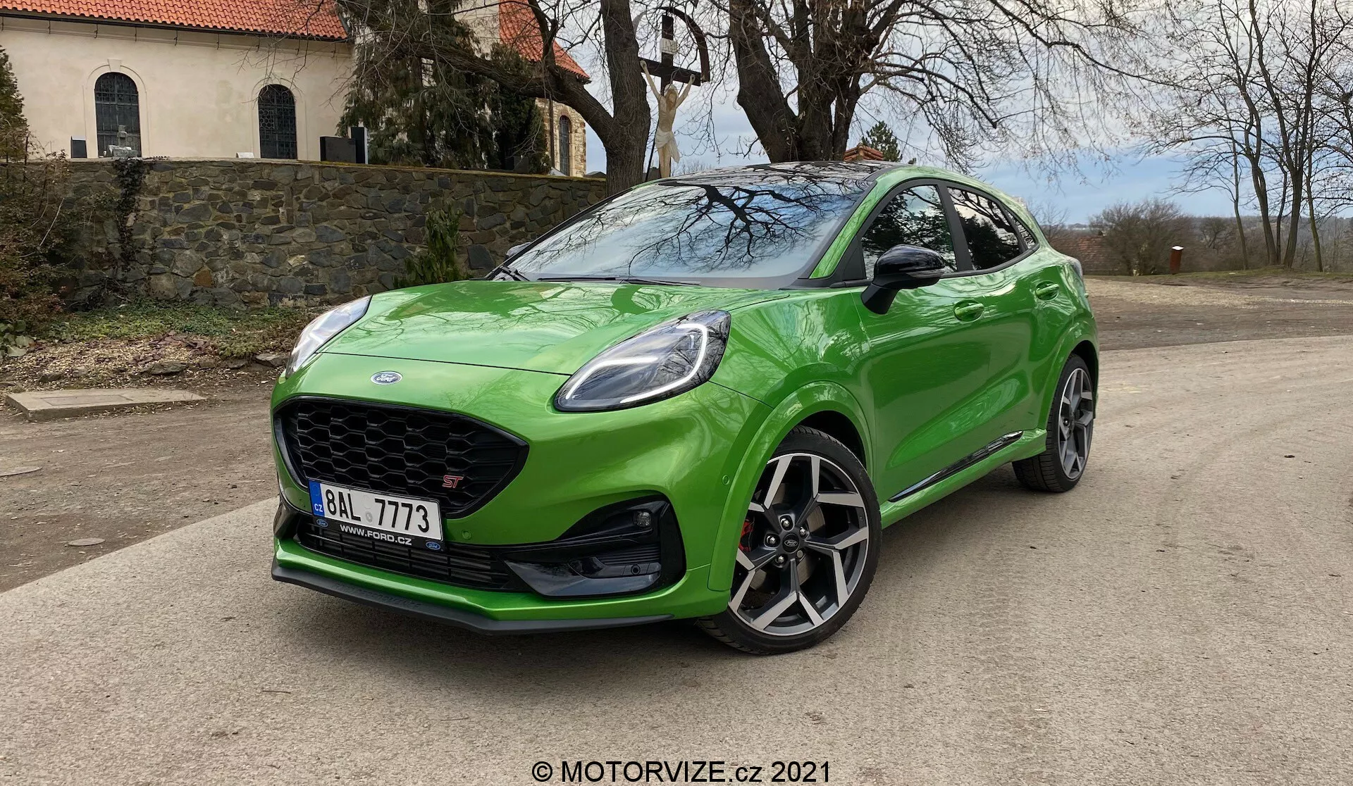Bilden visar en ljusgrön 2020 Ford Puma ST halvkombi parkerad på en belagd yta, sedd från en trekvarts främre vinkel. Bilen har en stor motorhuv med bikakemönster, aerodynamiskt utformad främre stötfångare med luftintag, skarpa LED- eller HID-strålkastare med DRL och en huv med dynamiska linjer. Förarens sidprofil framhäver stora lättmetallfälgar med flera ekrar, sidoskirtar och en kompakt, sportig siluett med sluttande taklinje. Vindrutan är lutande för en aerodynamisk profil med lätt tonade fönster. Bilens glansiga gröna lack och dagsljuset med mjuka skuggor under bilen bidrar till dess oklanderliga utseende. Logotyper på grillen och en främre nummerplåt är synliga, placerade mot en naturlig bakgrund med träd.