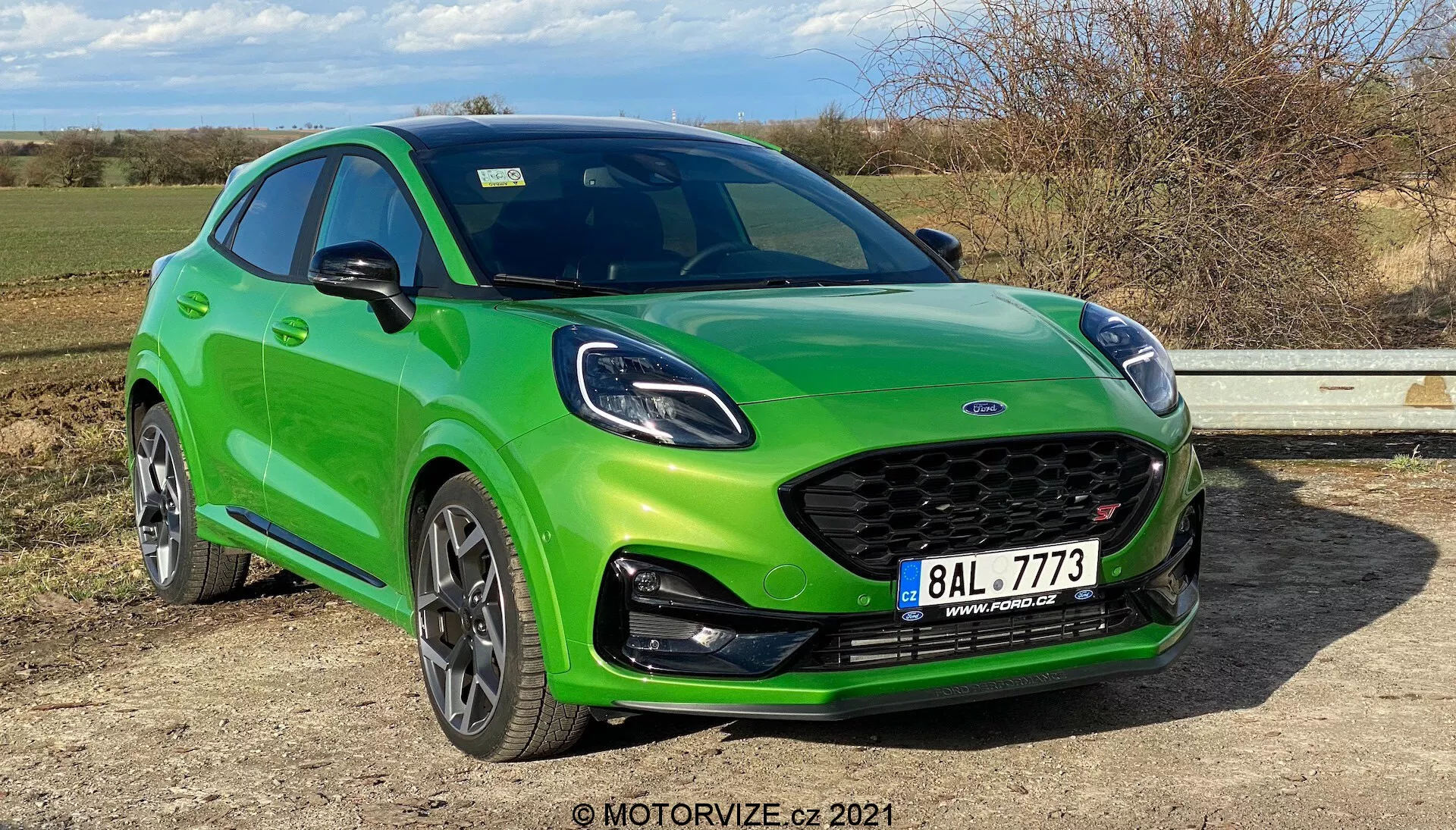 En livfull grön 2020 Ford Puma ST visad från en trekvarts fram-vänster vinkel, framhäver sin sidoprofil och frontdesign. Distinkt galler med tillverkarens emblem i mitten, sportig stötfångare med integrerade dimljus och en läppspoiler. Huven har sportiga konturer, smala strålkastare med LED-dagljus. Den dynamiska halvkombi-sidoprofilen har en sluttande taklinje, sidobackspeglar i karossfärg, svarta skyddslister längs dörrarnas nederkant och hjulhus, framträdande lättmetallfälgar troligen med däcktrycksövervakningssystem och synliga skivbromsar. Bilen är parkerad på en plan yta, med en glansig lack, som totalt sett betonar sin sportiga design, belysningsteknik och prestationsfunktioner i naturligt dagsljus.