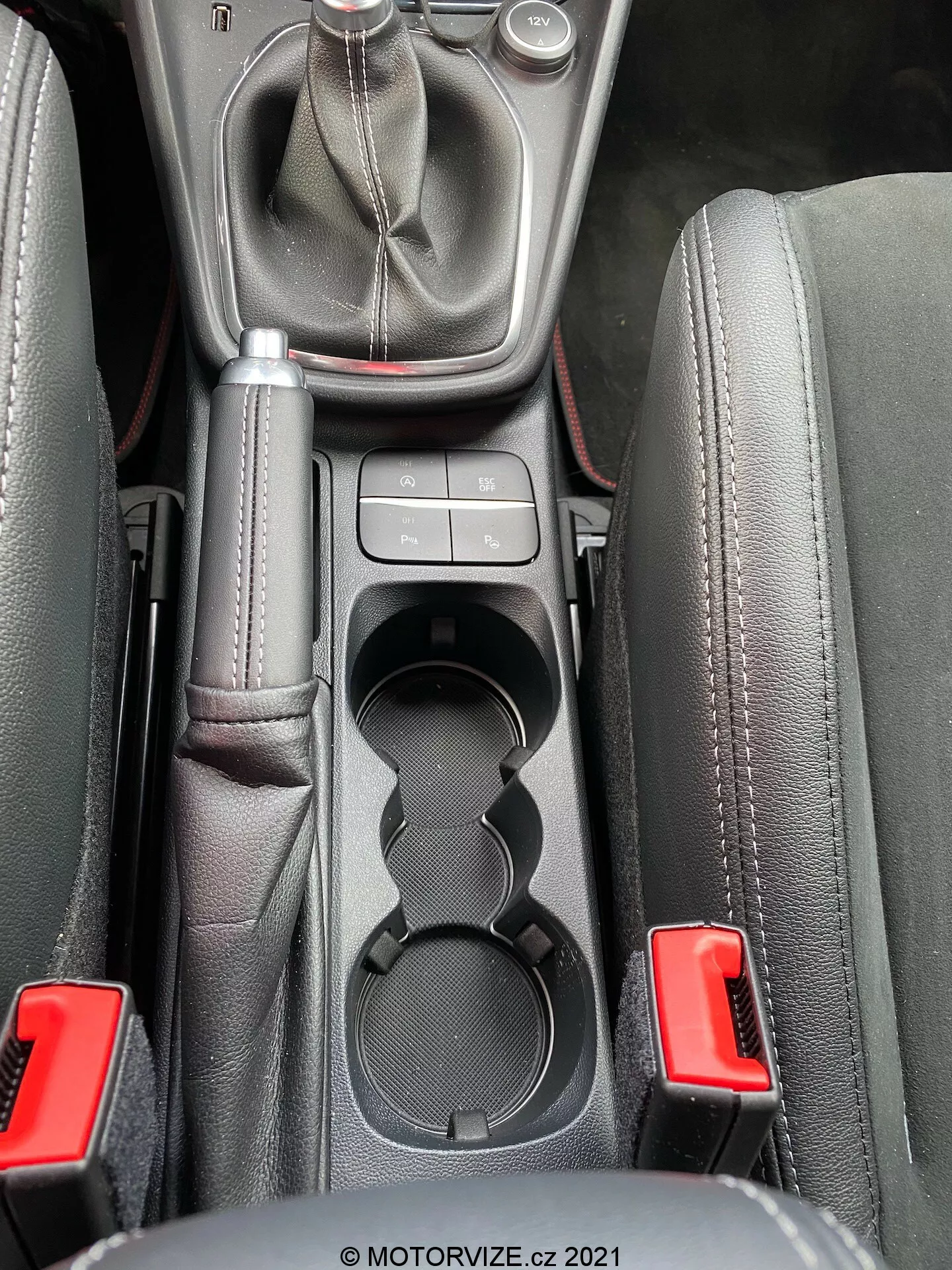 Närbild av interiören i 2020 Ford Puma ST:s mittkonsolområde taget från ögonhöjd, som visar en manuell växelspak med ett läderhölje och en metalltopp, omgiven av mittenkonsolens kontroller, dubbla mugghållare, ett vadderat armstöd och säkerhetsbälteslås i naturligt ljus.