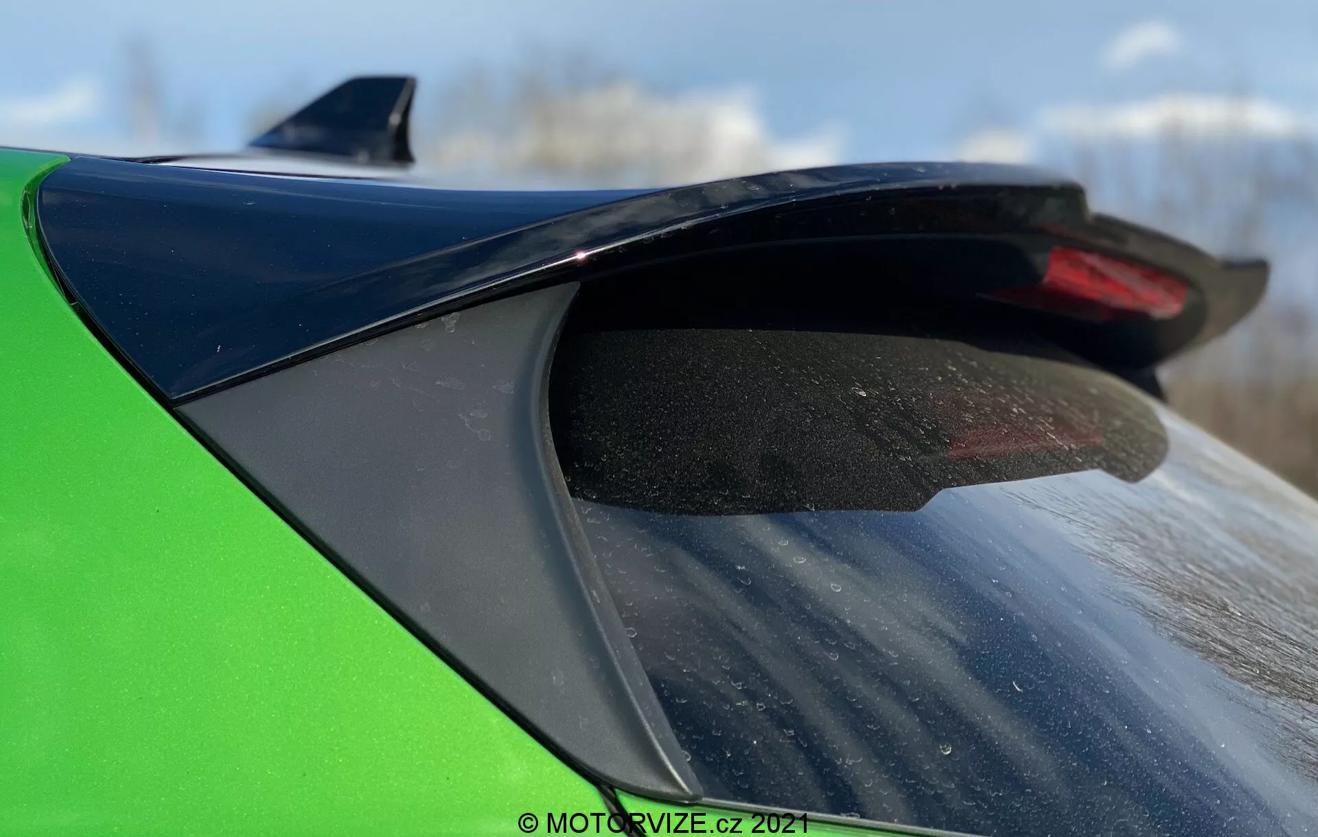Närbild av bakparten på en Ford Puma 2020, med fokus på svart glansig bakspoiler och grön bilkropp. Spoiler med damm och smuts, monterad på bagageluckan. Bakrutan med synliga defroster-linjer. Suddig utomhusbakgrund med starkt ljus och kort skärpedjup.
