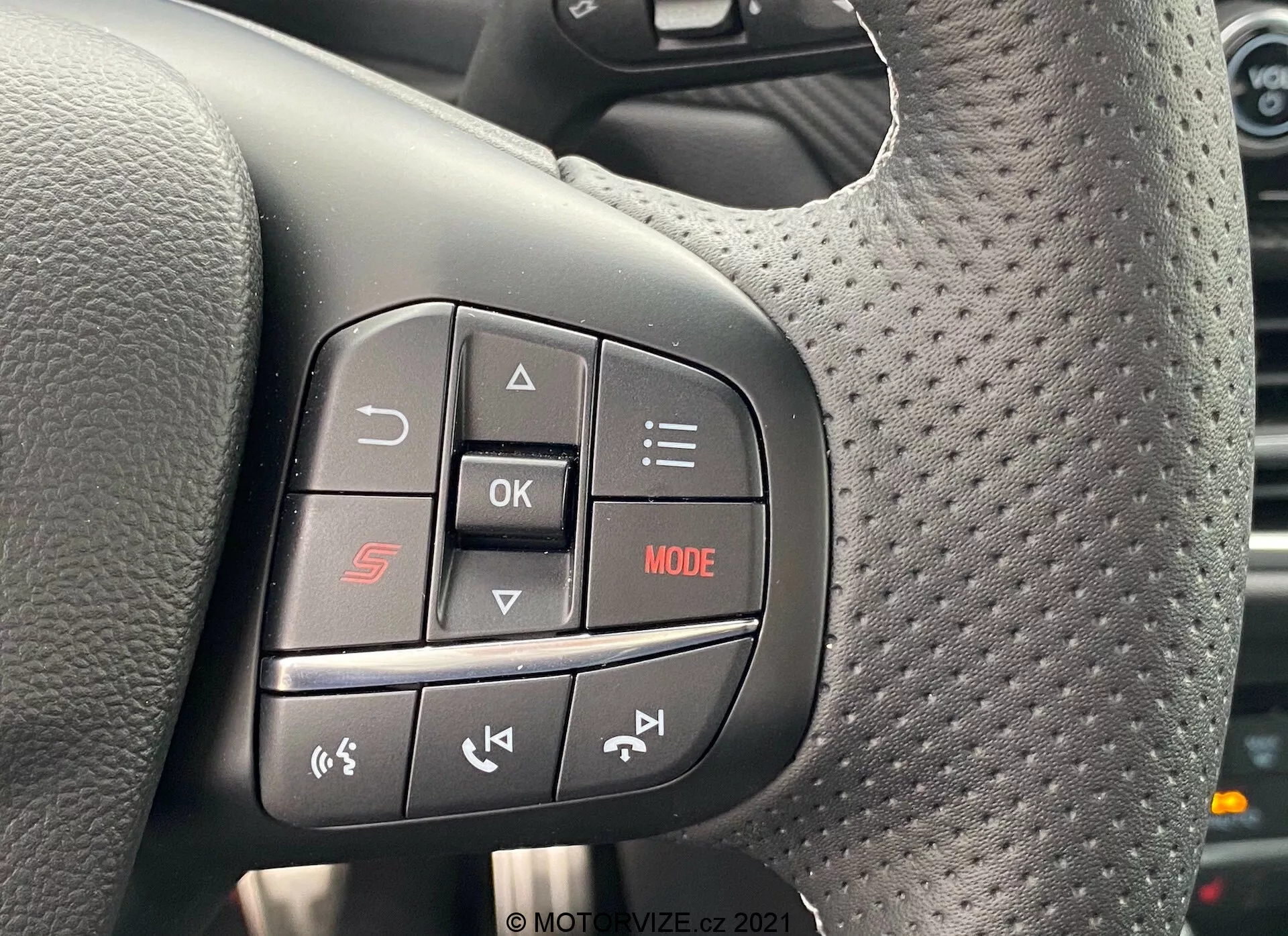 Närbild av de multifunktionella rattreglagen i Ford Puma 2020. Fokuset är på vänstersidans reglage, svarta knappar med röda och vita symboler. Överst 'S'-knappen för sportläge, mittre 'OK'-knappen, 'MODE' med röd LED, telefonikon under 'OK', och pilar för mediakontroll. Perforerat mönster runt kontrollområdet antyder en sportig eller exklusiv design.