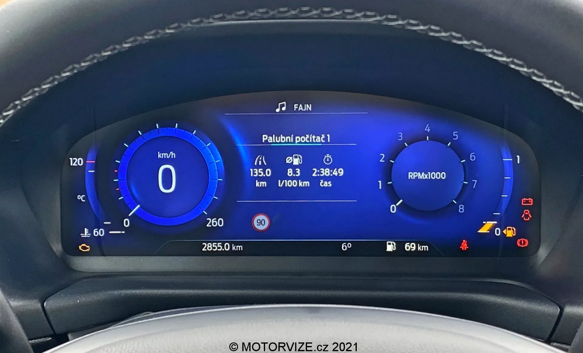 Närbild av den moderna digitala instrumentpanelen i en Ford Puma ST 2020 från förarens perspektiv. Bilden framhäver den livfulla blå digitala instrumentpanelen inramad av rattens kant. Panelen visar en hastighetsmätare, varvräknare, bränsleförbrukningsdata, total körsträcka, utomhustemperatur och olika varningssymboler, vilket illustrerar avancerade biltteknologier.