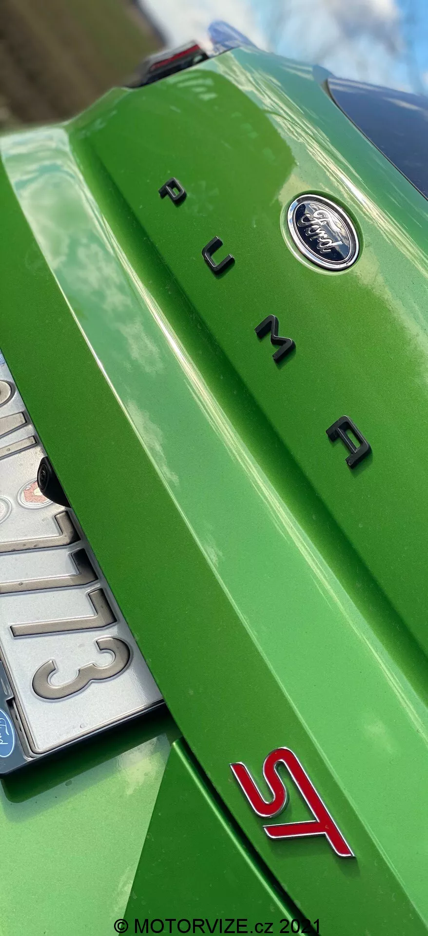 Närbild, låg vinkel bakifrån av en ljusgrön Ford Puma 2020. Synligt är en horisontell bakspoiler med ett centralt, cirkulärt emblem, kromat 'PUMA' text och en sportig röd 'ST' märkning, vilket indikerar sportmodellen av bilen. Bilens kaross reflekterar ljusa utomhusljusförhållanden.