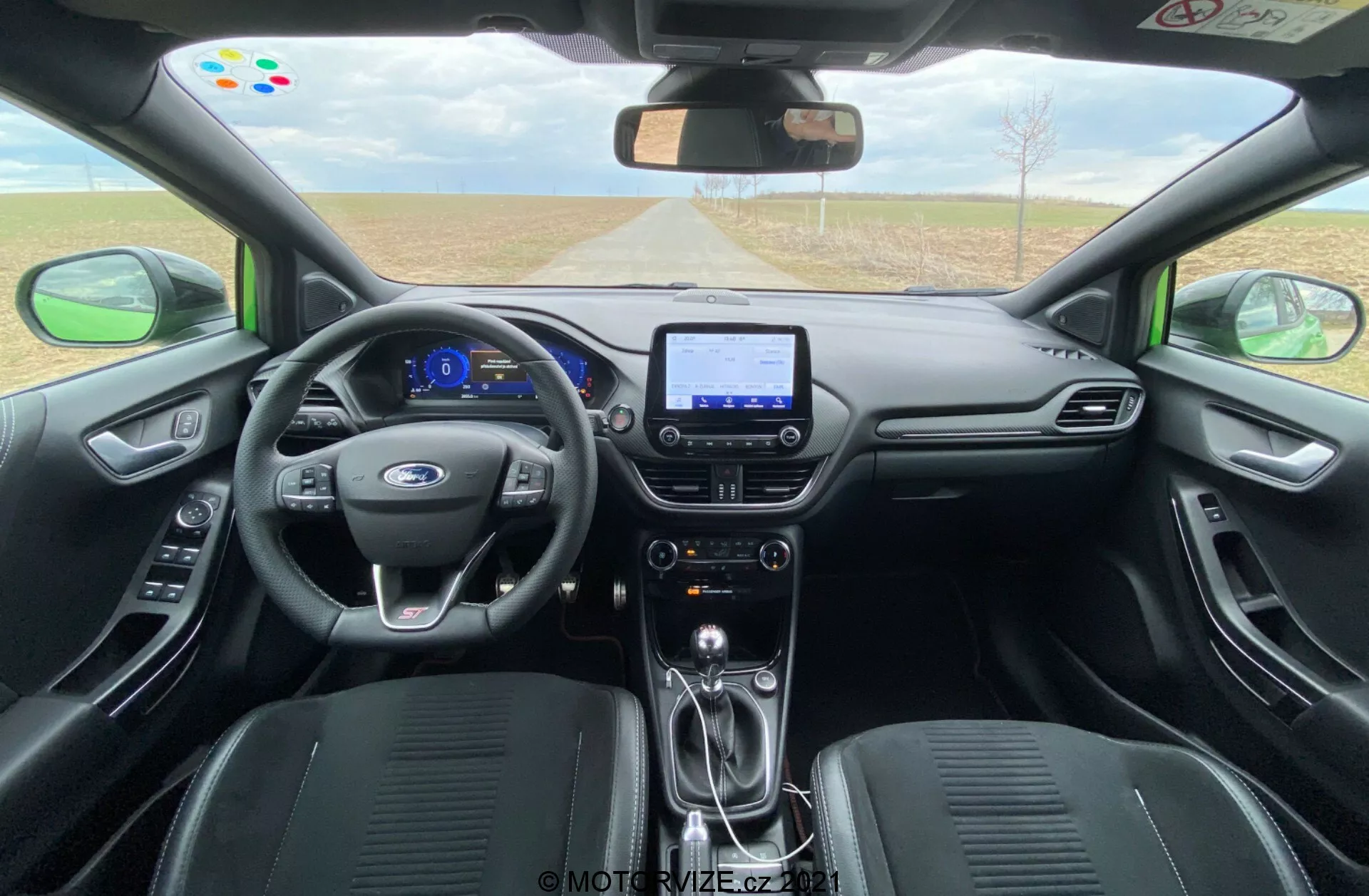 Interiören i en Ford Puma ST 2020 sedd från baksätet, med en treekrad läderklädd ratt med multifunktionskontroller, en digital instrumentpanel med blå upplysta displayer och en mittkonsol med en pekskärmsinfotainmentsystem som visar navigation. Funktioner inkluderar en synlig växelspak, elektronisk parkeringsbroms, elegant interiördesign med mörka material och svarta, skulpterade säten. Främre vindrutan erbjuder en klar, vidsträckt utsikt över vägen framför, medan dörrarna har kontroller för elhissar och speglar.