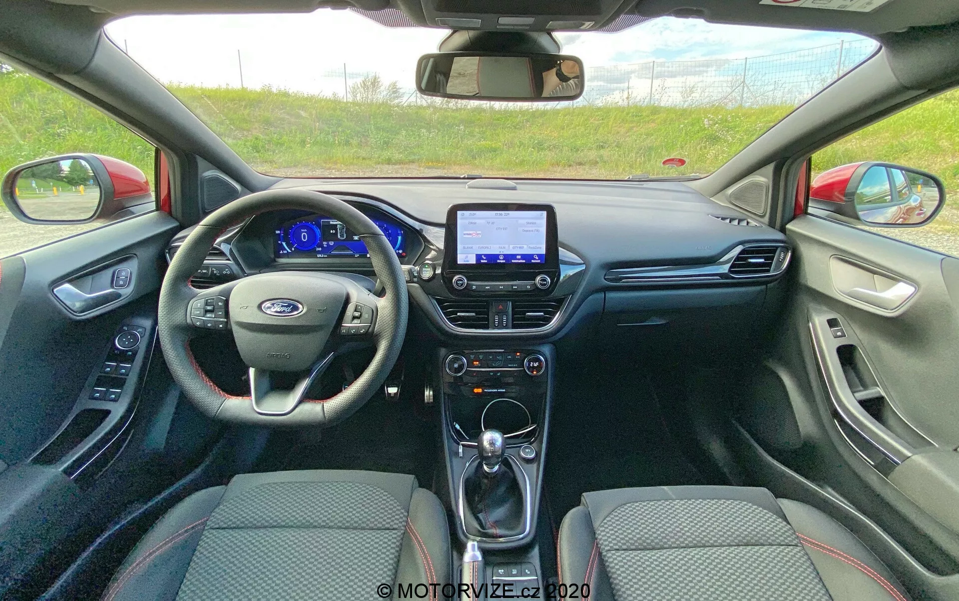 Interiörvy av en Ford Puma 2020 från baksätet som tittar framåt. Bilden visar hela instrumentpanelen och en del av framsätena, med fokus på förarsidan. En centralt placerad ratt visar tillverkarens logotyp och symmetriska kontrollknappar för media, farthållare och handsfree-kommunikation. Den digitala instrumentgruppen inkluderar en hastighetsmätare, varvräknare, bränslemätare och olika informationsdisplayer. I mitten av instrumentpanelen syns en infotainment-pekskärm, vilket indikerar funktioner som navigation, fordonsinställningar och multimediafunktioner. Under pekskärmen finns manuella klimatkontrollvred och knappar, växelspak för automatlåda, USB-portar och ett 12V-uttag. Interiören är väl underhållen med en tvåfärgad tyguppsättning på sätena och funktionella dörrpaneler med fönsterkontroller och högtalargaller. Backspegeln och en sensor- eller kameramodul för ett avancerat förarassistanssystem är också synliga, vilket förbättrar säkerheten och förarens bekvämlighet.