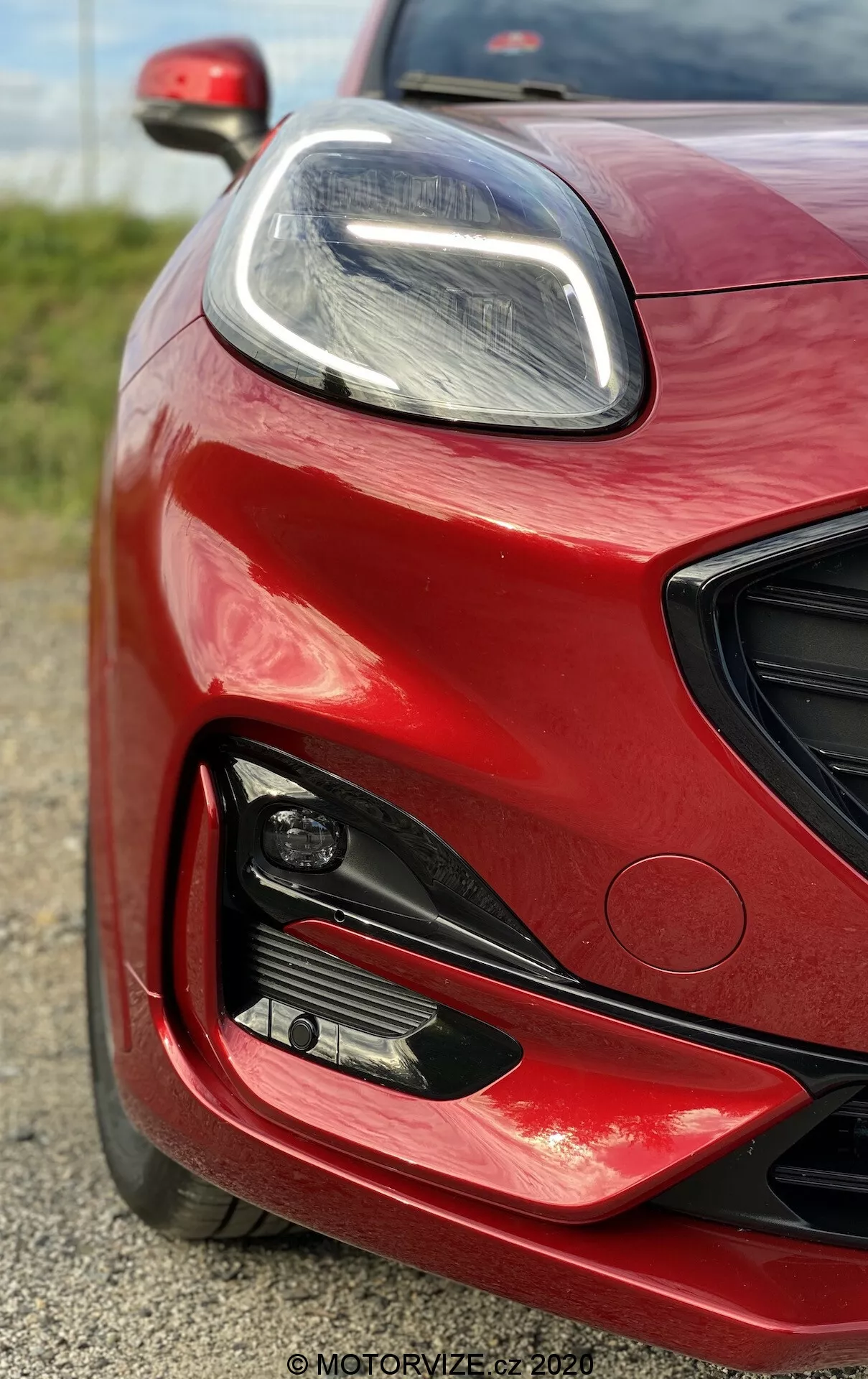 Närbild på den vänstra främre delen av en 2020 Ford Puma i livfull röd med glansig finish, uppvisar elegant LED-strålkastare, stora luftintag och dimljus, som betonar dess moderna, prestandainriktade design.