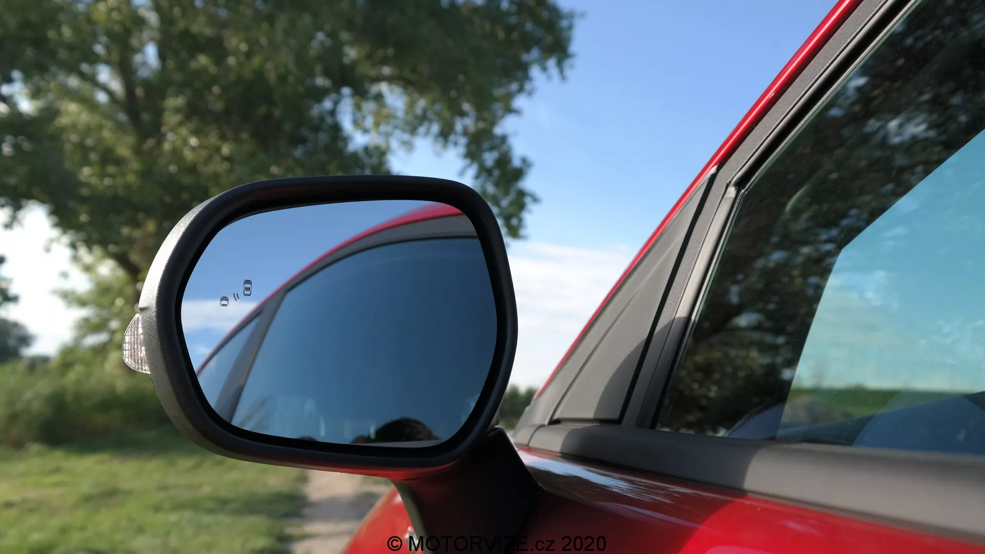 Närbild av sidospegeln på en röd Ford Puma 2020, med fokus på den aerodynamiska svarta spegelhuset med en traditionell platt spegel och en döda vinkeln-sektion. Bilden visar förarsidans dörr och bakre kvartspanel i dagsljus, vilket framhäver bilens kurvor och designlinjer. Scenen antyder att fordonet är parkerat i ett öppet område med naturliga omgivningar.