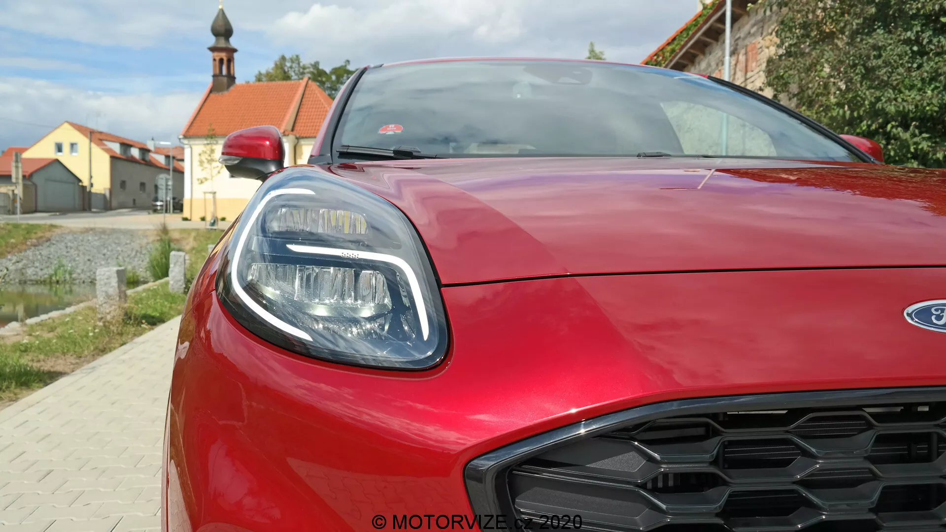 Närbild av fronten på en röd Ford Puma 2020, som visar den högra strålkastaren med eleganta LED-element, den skulpturala aerodynamiska motorhuven och bikakemönstrade grillen. Bilden är tagen från en låg vinkel och fångar bilens moderna stil och sportiga detaljer.