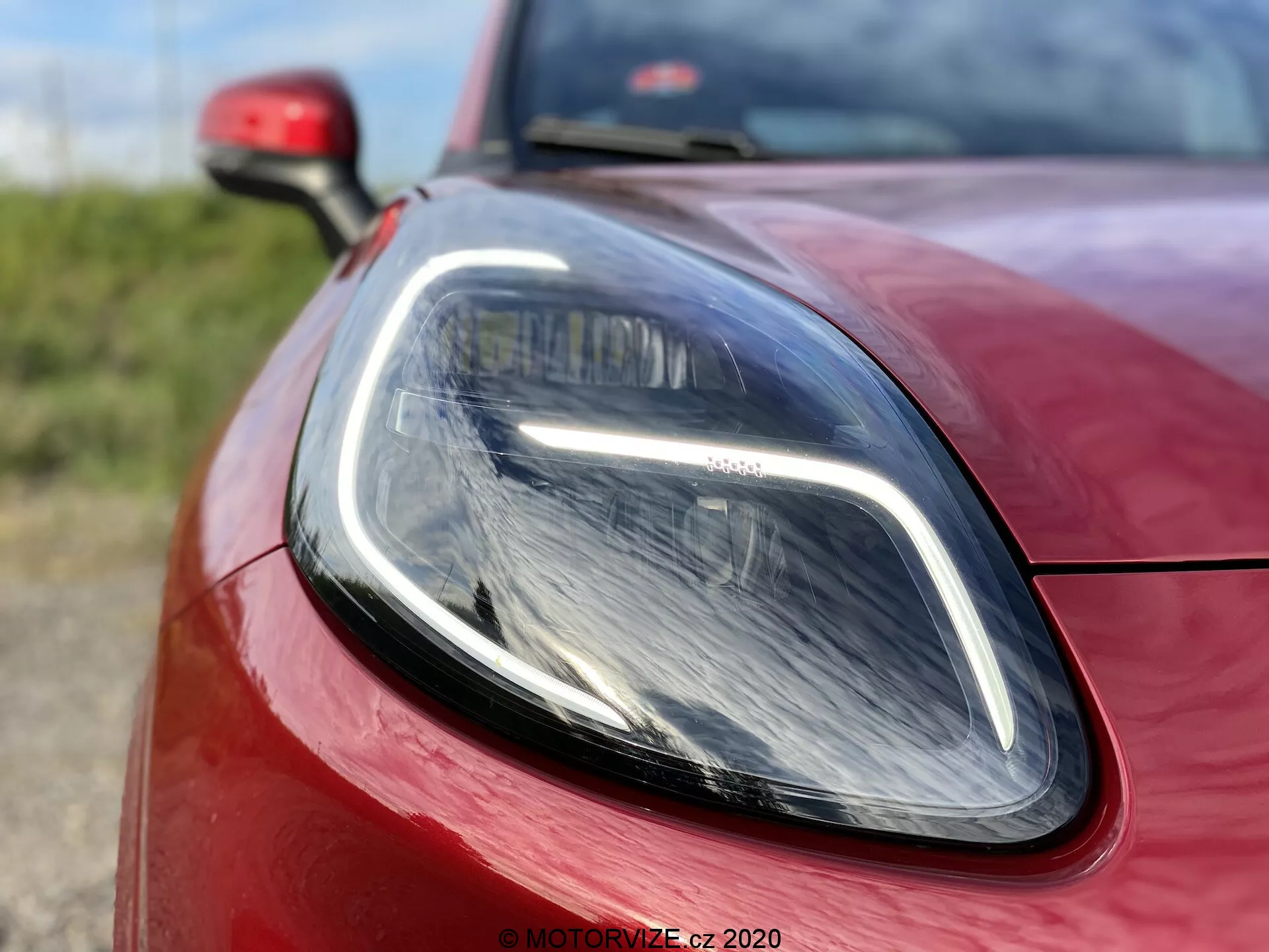 Närbild av 2020 Ford Puma, med fokus på en sektion av strålkastaren på förarsidan och den omgivande karossen. Bilden är tagen från en låg, något förskjuten vinkel och framhäver de eleganta LED-dagsljusen och den moderna projektorstrålkastaren. Den livfulla röda färgen och de exakta paneljusteringarna betonar bilens sportiga, aerodynamiska design.