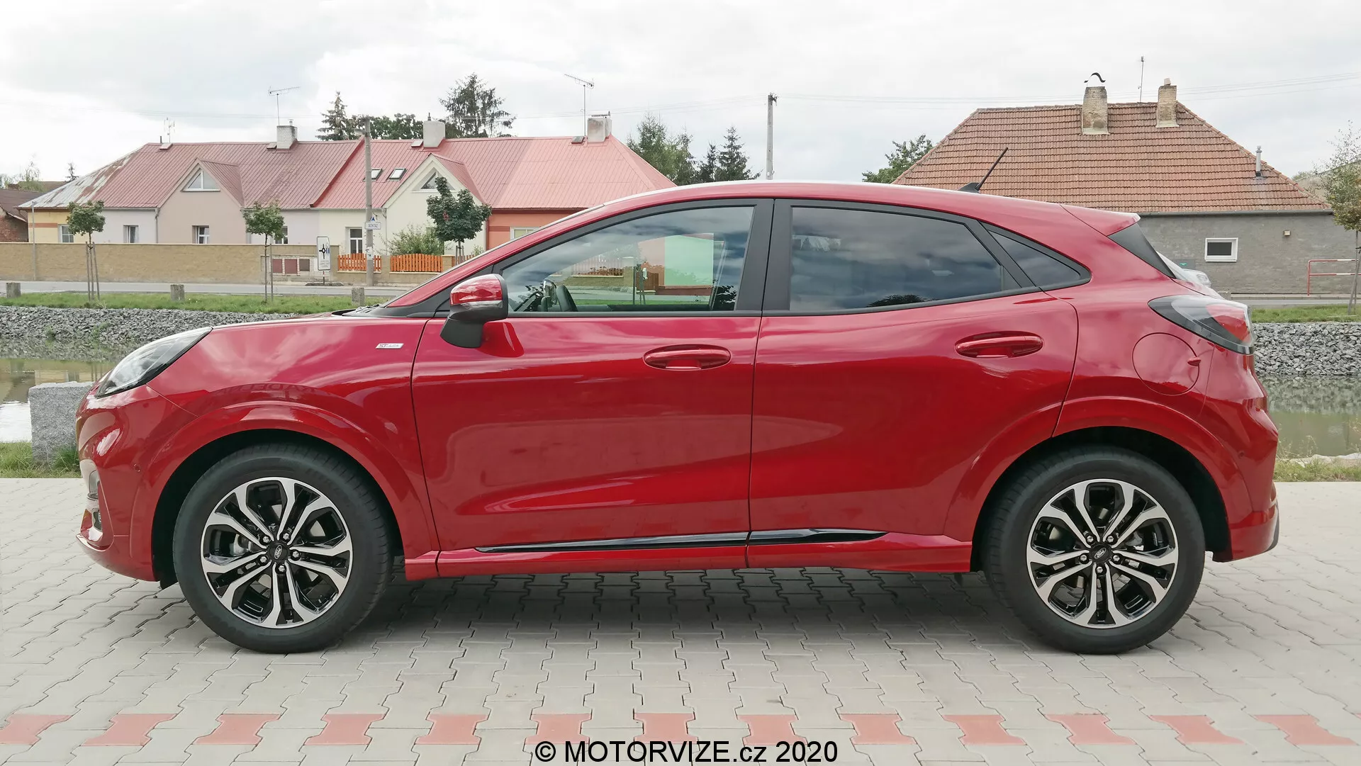 Sidoprofilvy av en röd fyrdörrars 2020 Ford Puma hatchback, tagen under dagen på en parkeringsplats med gräs och bostadshus i bakgrunden. Bilen har aerodynamiska linjer, flerspåriga lättmetallfälgar, färganpassade sidospeglar och dörrhandtag samt en nedåtgående taklinje. Avsaknaden av synliga nyckelhål antyder ett möjligt nyckelfritt system.