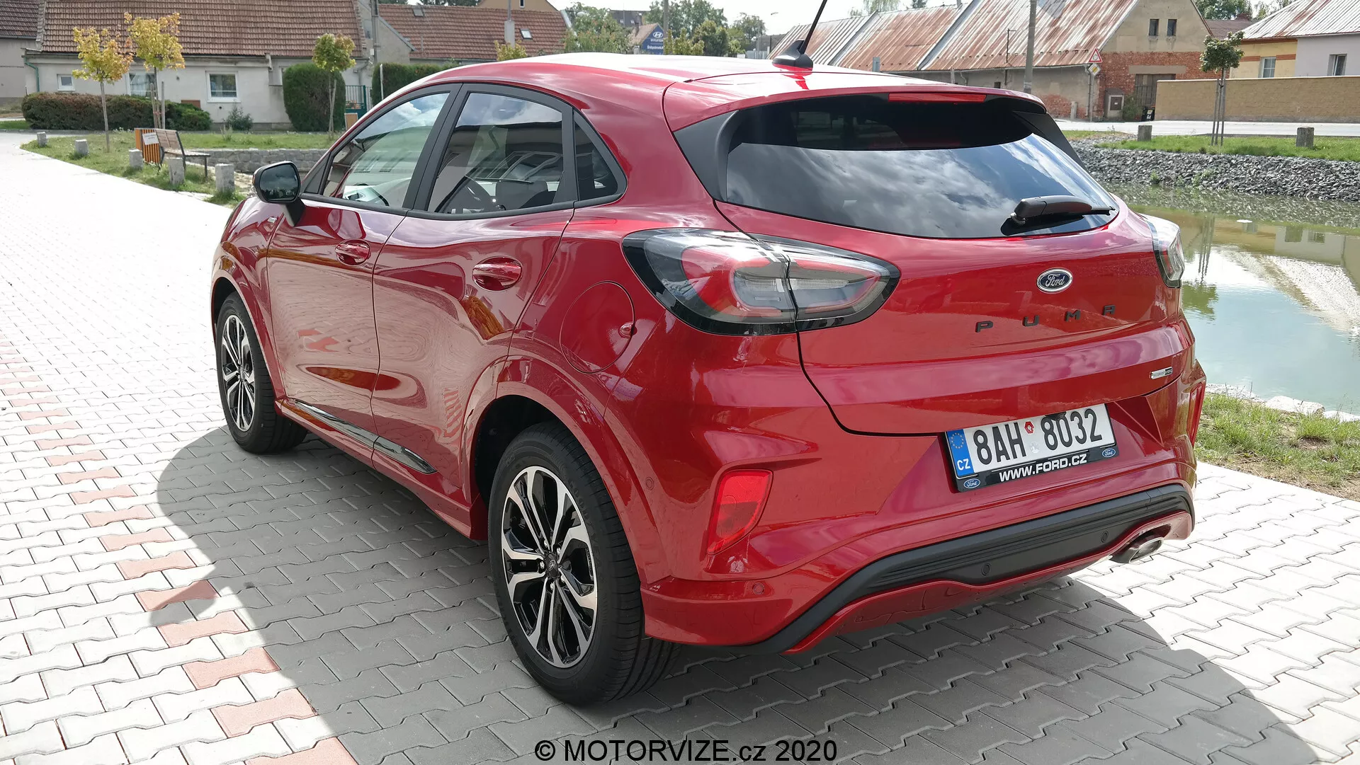 Tre-kvarts bakre vy av en modern Ford Puma 2020 i livfull röd färg, fotograferad i dagsljus. Bilden framhäver bilens aerodynamiska konturer, de karakteristiska baklamporna som sveper runt sidorna och de integrerade sensorerna i den bakre stötfångaren, vilket antyder parkeringsassistansfunktioner. Dubbla avgasrör och en bakre spoiler förstärker dess sportiga utseende, medan de flerspända fälgarna och karossfärgade elementen som sidospeglar och dörrhandtag bidrar till dess moderna design. Bakre vindrutetorkare synlig, vilket indikerar praktiskhet under ogynnsamma väderförhållanden.