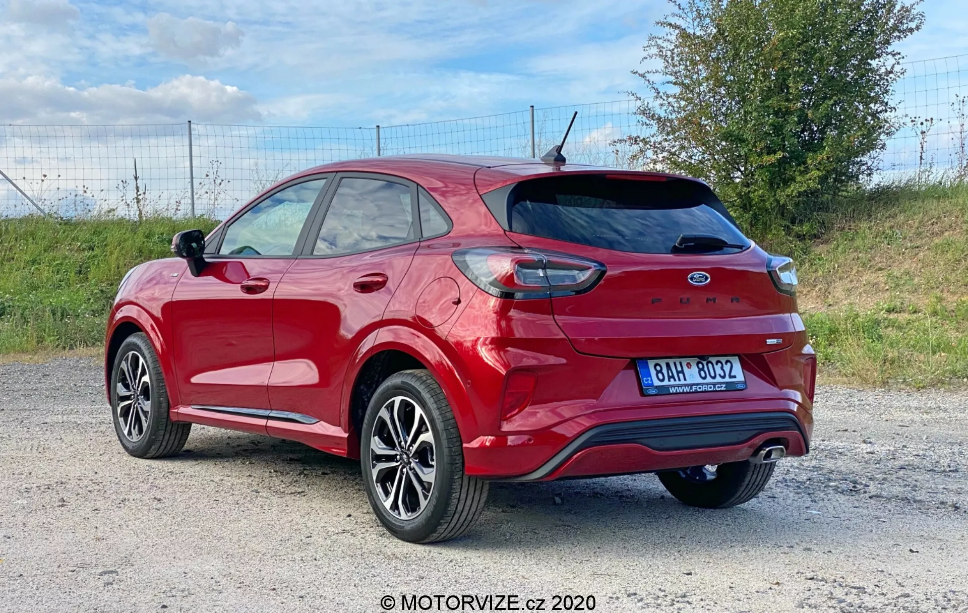 Bilden visar en röd Ford Puma 2020 crossover/SUV fotograferad från en tredjedel bakvinkel, vilket visar bakhjulet och däcket, en del av framdörren och bakdörren för passagerare, bakre kvarts panelen och hela bagageluckan med integrerad spoiler och bakre vindrutetorkare. Synliga egenskaper inkluderar bakljus som delvis sveper runt bakskärmen, bakre stötfångare med reflexer och parkeringssensorer, dubbelt avgasystem, hajfeneantenn, bakrutan med avfrostningslinjer, aluminiumfälgar med multi-ekrade design och lågprofildäck, tonade rutor, takräcken och en sidospelskåpa som matchar bilens färg. Fordonet står i en utomhusmiljö, sannolikt på en grus- eller grusväg, med ett gräsområde synligt i bakgrunden vilket tyder på en lantlig eller halv-lantlig miljö.