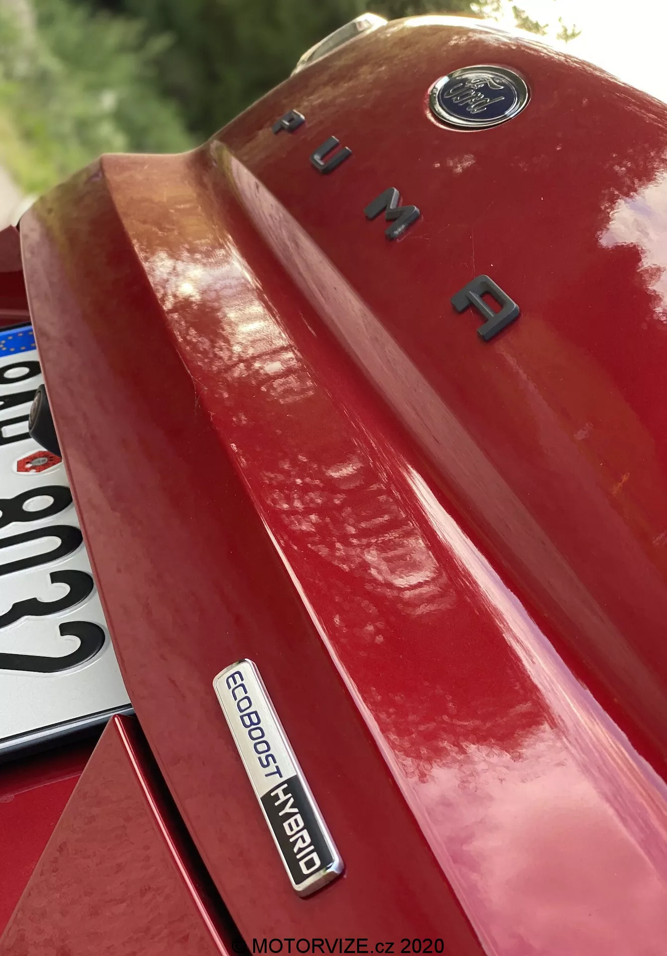 Närbild av vänster bakre kvarts av en röd Ford Puma 2020, tagen från en uppåtvinklad vinkel, som tydligt visar märken och branding. Bilden framhäver Ford-emblemet högst upp, modellnamnet 'PUMA' i kromade bokstäver på bagageluckan och 'ECOBOOST HYBRID'-märket, vilket anger avancerad hybridteknik. En del av registreringsskylten är delvis synlig nere till vänster. Fordlacke reflekterar ljus och framhäver bilens design och tekniska sofistikering.