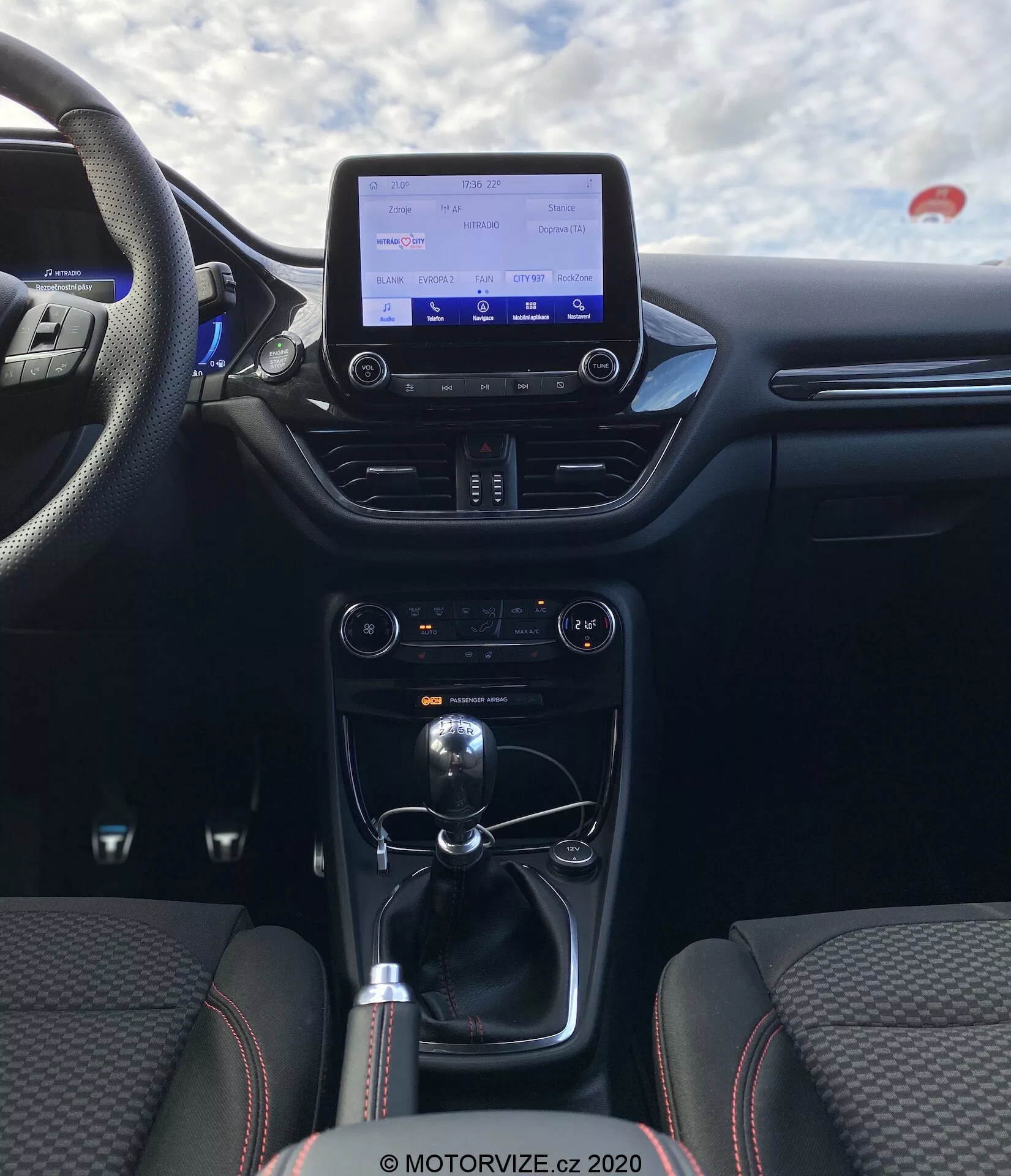 Interiörvy av 2020 Ford Puma från bakre passagerarens perspektiv med fokus på centrala cockpitområdet. Bilden lyfter fram mittkonsolen och instrumentpanelen, inklusive en infotainmentskärm, klimatanläggning, växelspak för manuell växellåda och anslutningsalternativ som USB-portar och 12V-uttag. Sätena är klädda i tyg med ett distinkt sömmönster och sido-stödd, betonar komfort och sportighet.