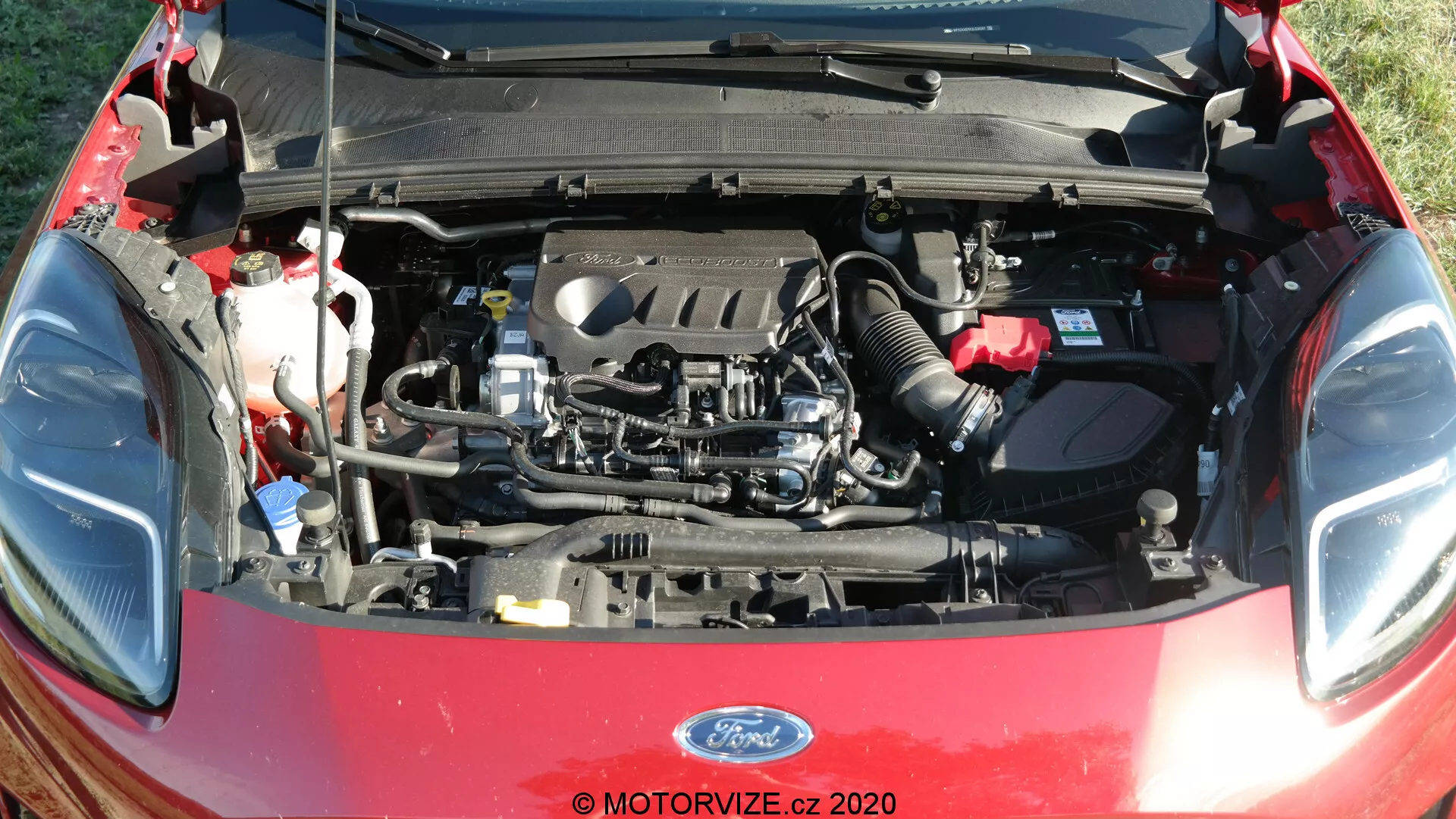 Detaljerad vy av Ford Puma 2020 motorutrymme med den öppna huven, som visar motorkomponenter inklusive motorblock, ventilkåpa, insugningsgrenrör och avgasgrenrör. Synliga element inkluderar också vätskebehållare, slangar, kablar, batteri, luftkanaler och delen av den öppna huven med isolering.