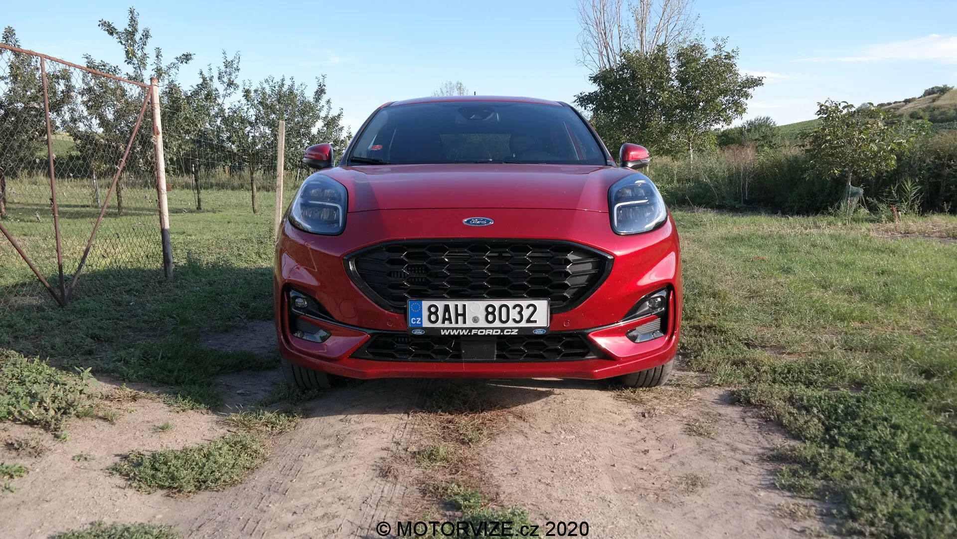 Frontal vy av en 2020 Ford Puma som visar dess framträdande nätgrill, eleganta LED-strålkastare och aerodynamiska motorhuvkonturer. Bilen är placerad på torr jord med gräsfläckar, vilket antyder en lantlig miljö.