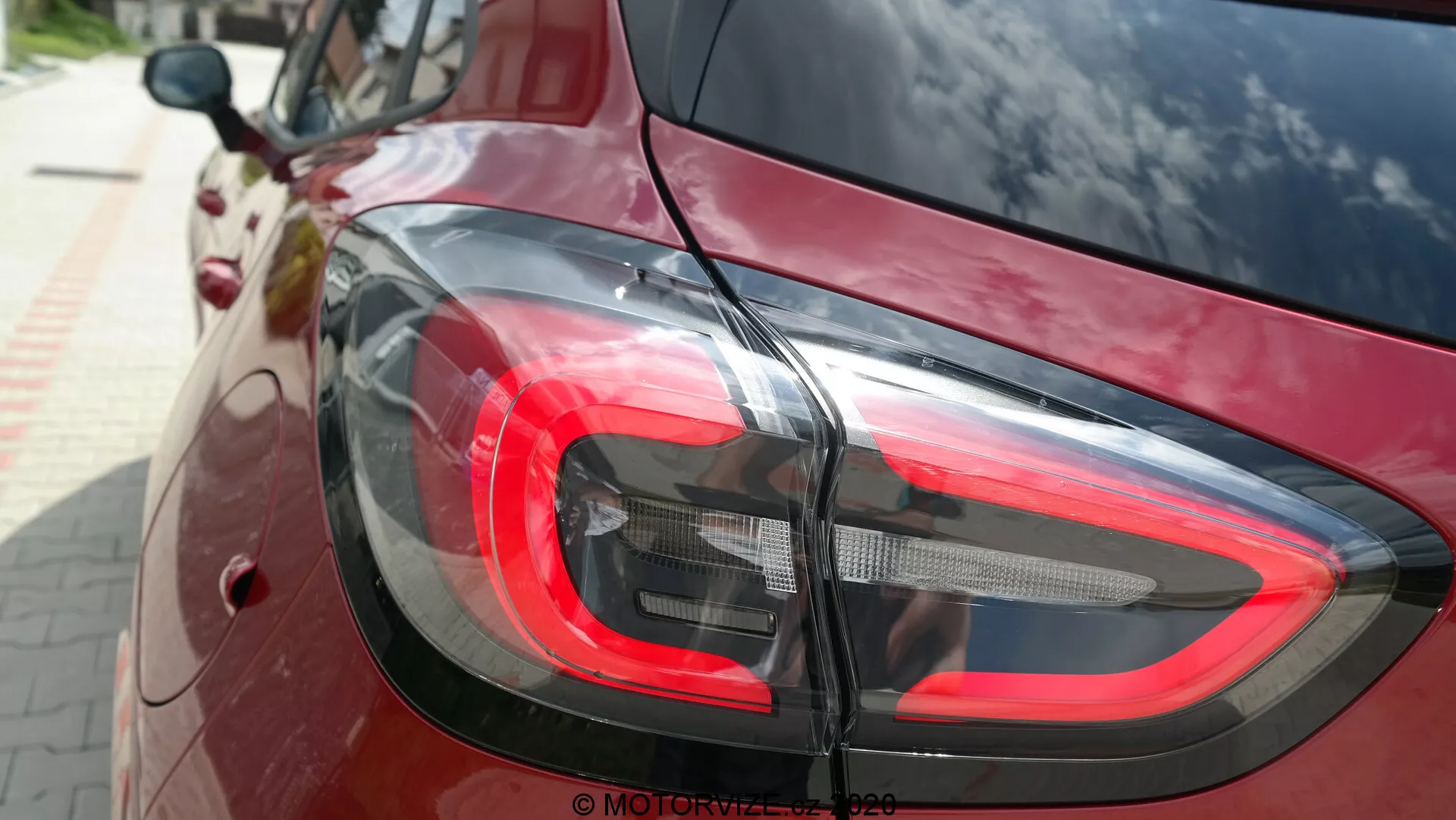 Närbild av den bakre delen av en Ford Puma 2020, med fokus på den moderna LED-bakljusdesignen från en något upphöjd vinkel. Det röda bromsljuset, de klara och mörkare segmenten för backljus och blinkers smälter sömlöst in i bilens djup röda eller vinröda högblanka lack som speglar omgivningen. Bilden framhäver bilens kurviga och moderna stil samt dess funktionella belysningsteknik.