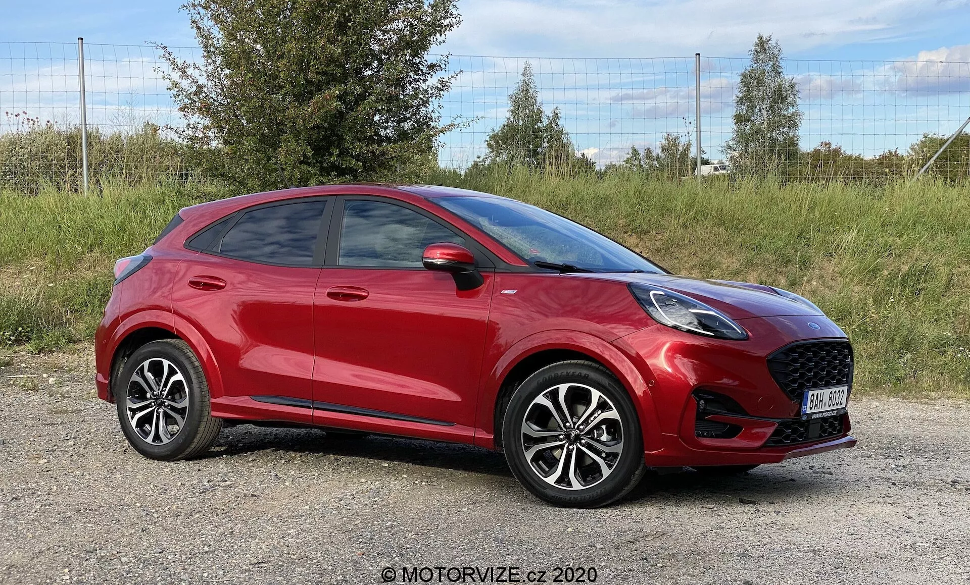 Professionellt fotografi av en röd 2020 Ford Puma kompakt crossover SUV, placerad på ett gräsbevuxet område med lövverk i bakgrunden. Den främre sidovinkeln framhäver den framträdande frontgrillen med bikakemönster, eleganta LED-strålkastare och delvis synlig sidobackspegel. Den sportiga, sluttande taklinjen, distinkta dörrlinjer, tydligt synliga handtag, lättmetallfälgar med flerradigt mönster och lågprofilsdäck bidrar till bilens moderna estetik. Bilden, tagen i nivå med bilen, fångar hela dess längd och framhäver dess samtida design.