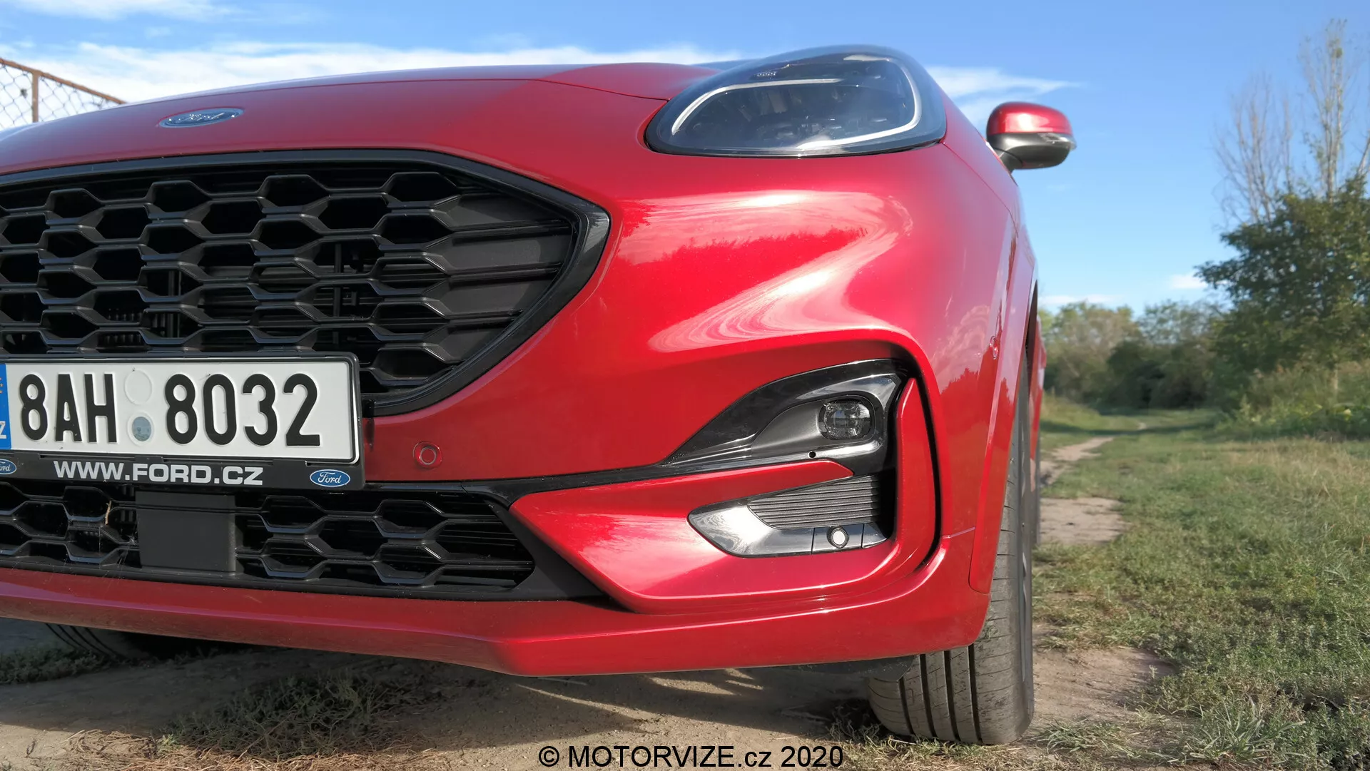 Närbild i vinkel av en röd 2020 Ford Puma med fokus på vänster framsida. Funktioner inkluderar honungskaksgrill, elegant LED/HID-strålkastare, främre stödfångare och skulpterad aerodynamisk motorhuv. En europeisk registreringsskylt med en tjeckisk webbplats är synlig. Moderna element som integrerade blinkers i sidospegeln är delvis synliga. Bilden, tagen från låg vinkel, använder naturligt dagsljus och kort skärpedjup för att framhäva bilens design i en utomhusmiljö.
