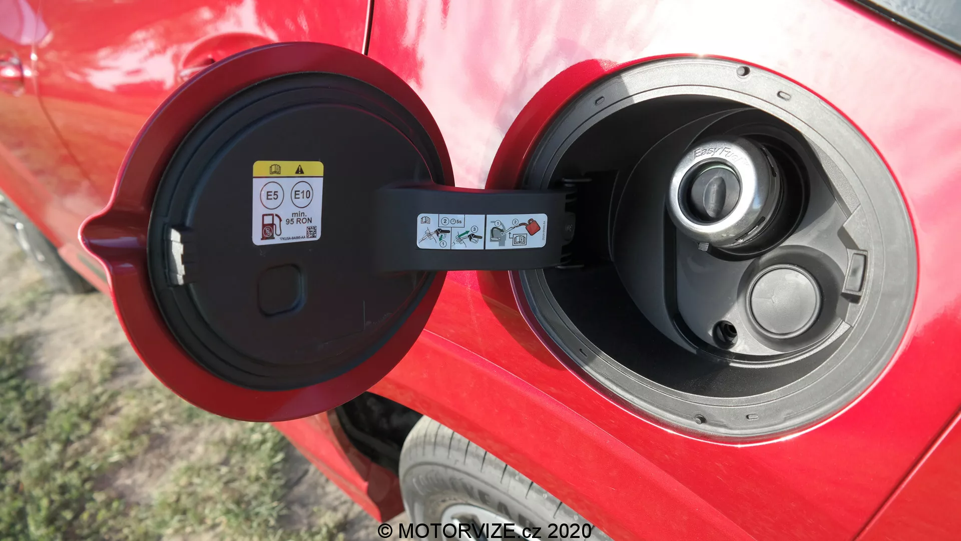 Närbild av en öppen Type 2-laddport på ett klarrött elektriskt eller plug-in hybridfordon, med fokus på det glansiga portområdet, gångjärnslocket och en informationsdekal synlig till vänster inuti portdörren, vilket antyder en väl underhållen bil med avancerad teknik för elfordon utomhus.