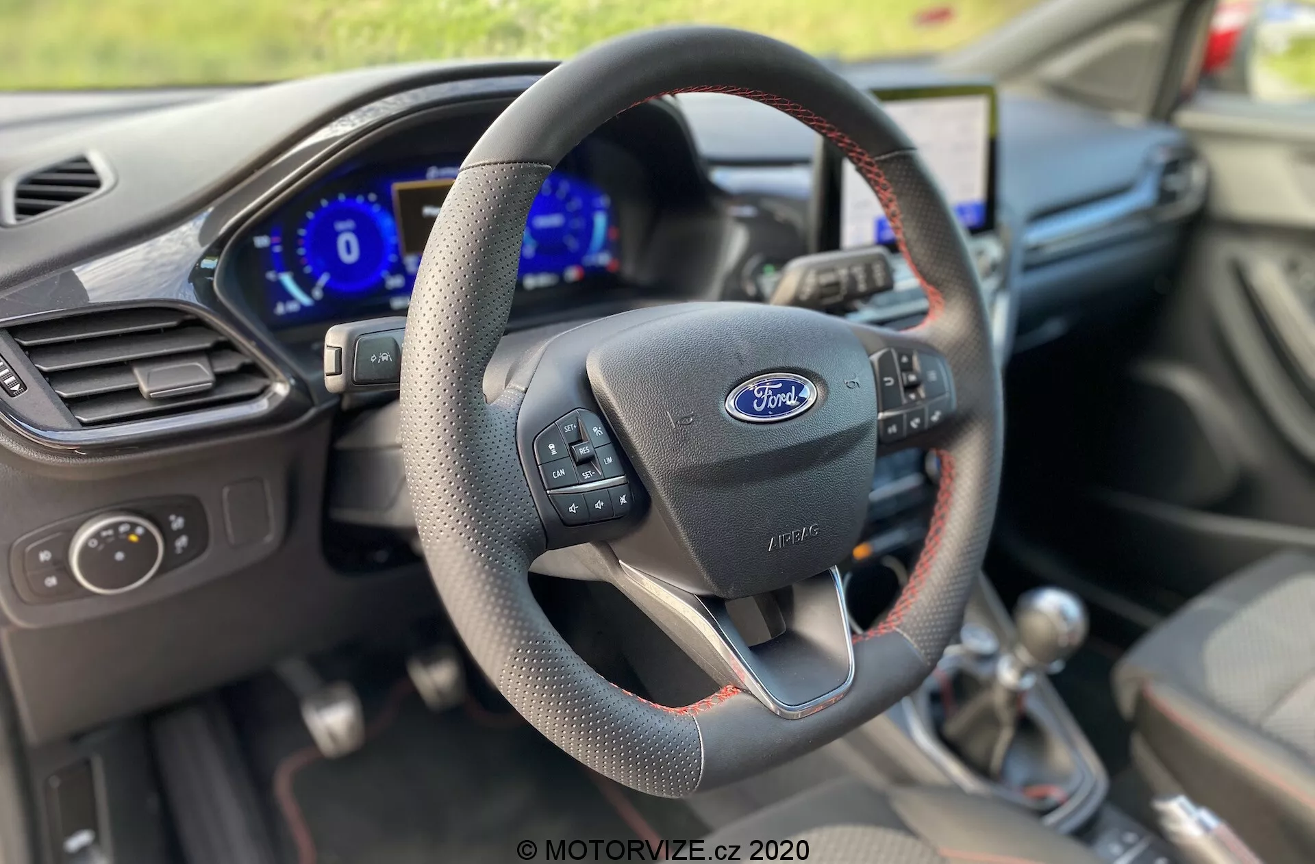 Interiör av en Ford Puma 2020 från förarens perspektiv, som framhäver en treekrad läderklädd ratt med ergonomiska grepp, multifunktionskontroller för infotainment och farthållare, och en framträdande Ford-logga. Instrumentbrädan bakom är digital och lyser blått, vilket indikerar att fordonet är igång. En manuell växelspak är synlig på mittkonsolen, vilket tyder på en mer engagerande körupplevelse. Förarens dörr visar kontroller för elhissar och speglar. Interiördesignen är funktionell, inte lyxig, med fokus på förarens interaktion med teknik och kontroller.