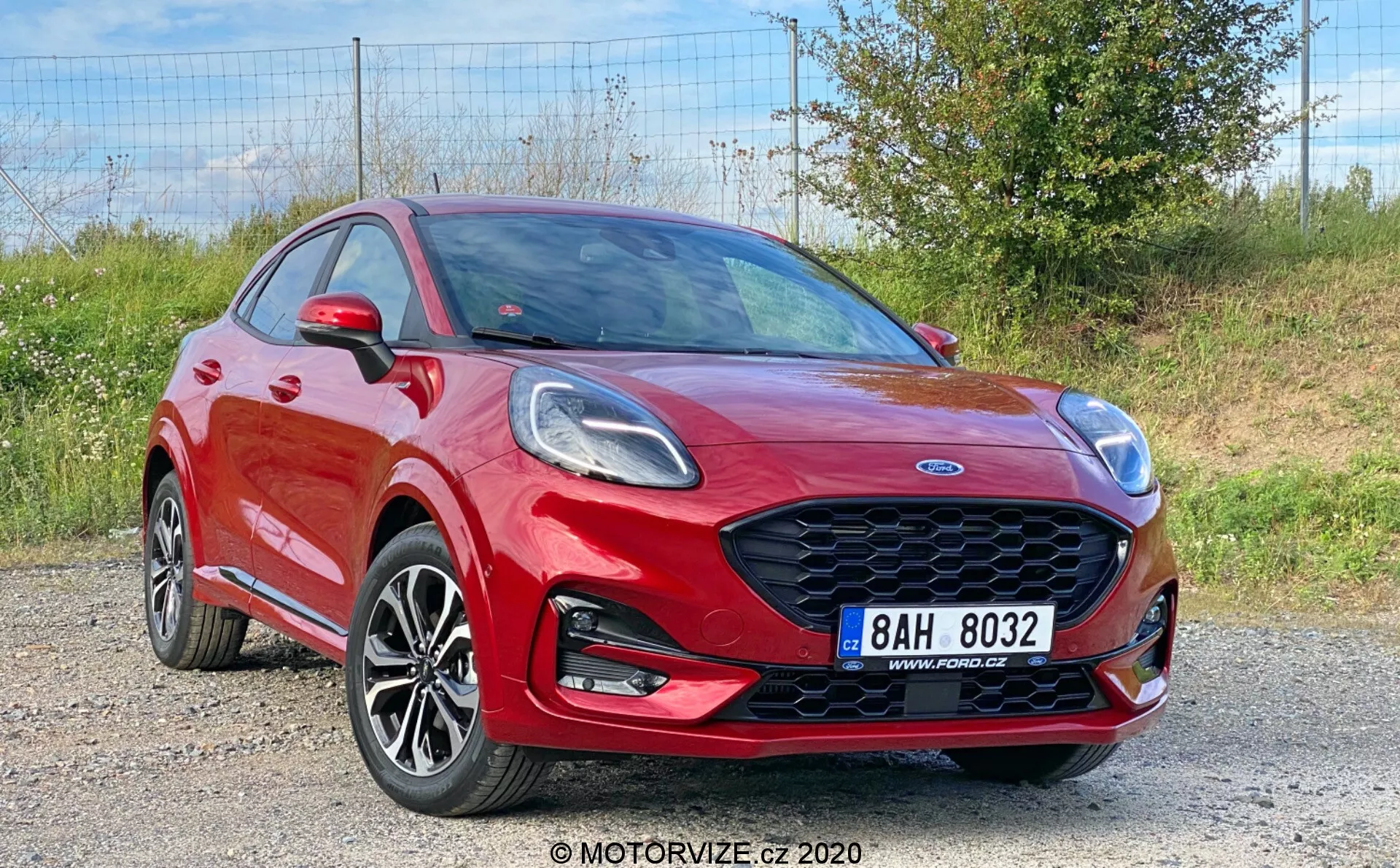 Bilden visar en livfull röd Ford Puma 2020, en crossover-fordon fångad från en främre trekvartsvy, som visar upp den eleganta främre grillen med bikupemönster, de vinklade strålkastarna, de flerspokrade legeringshjulen, den främre stötfångaren med integrerade dimljus och den markerade skulderlinjen. Den dynamiska motorhuven med veck, speglarna med integrerade blinkers och en del av både de främre och bakre dörrarna är synliga, vilket indikerar en fyr- eller femdörrars konfiguration, mot en grusbakgrund med växtlighet.
