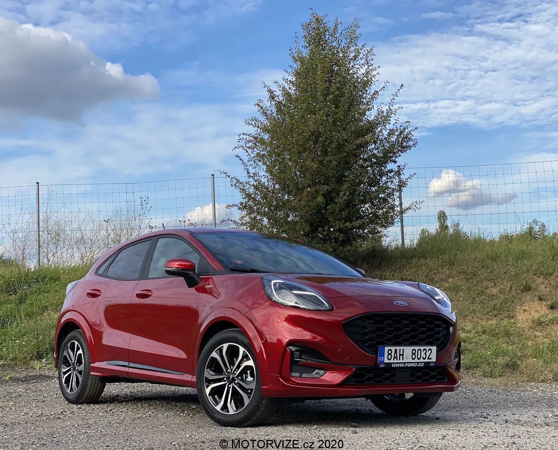 En dynamisk trekvart framifrån vy av en röd Ford Puma 2020 hatchback, som framhäver dess sportiga design, stora frontgrill med tillverkarens logotyp, eleganta LED- eller HID-strålkastare, dimljus integrerade i främre stötfångaren och flerspoked fälg i lättmetall. Bilen är placerad utomhus på en grusväg, med gräs, ett träd och ett staket i bakgrunden under en delvis molnig himmel.
