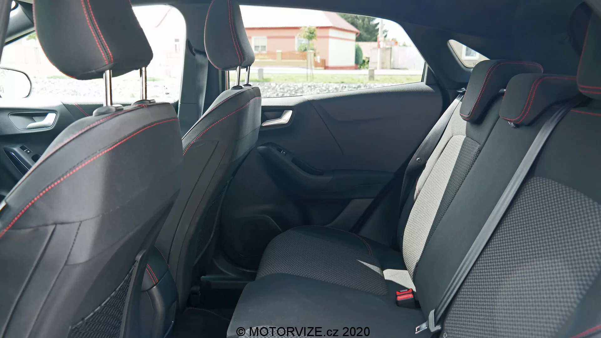 Interiörbild av Ford Puma 2020 med fokus på baksätena, tagen från utsidan av bakdörren på passagerarsidan, som visar den rymliga, sportiga designen med tyg- och lädermaterial, accentuerad med röda sömmar. Synliga funktioner inkluderar justerbara nackstöd, trepunktsbälten och delbara baksäten för ökad mångsidighet. Dörrpaneler med handtag, elektriska fönsterreglage och förvaringsutrymmen är också synliga, vilket framhäver ergonomiska och bekvämlighetsaspekter.